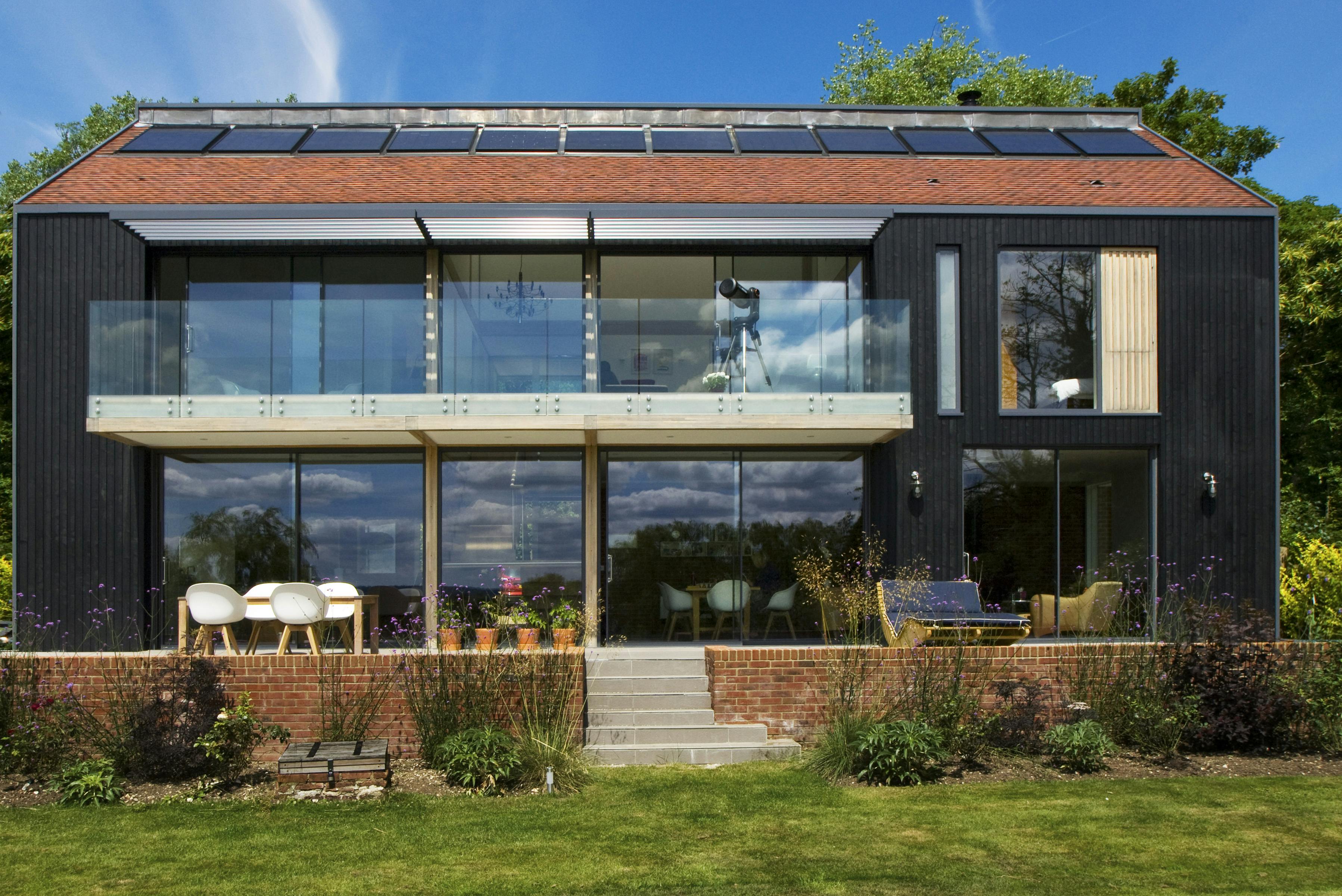 Large glazed windows and doors of the back of a large oak glulam framed house with a raised patio and green lawn in front