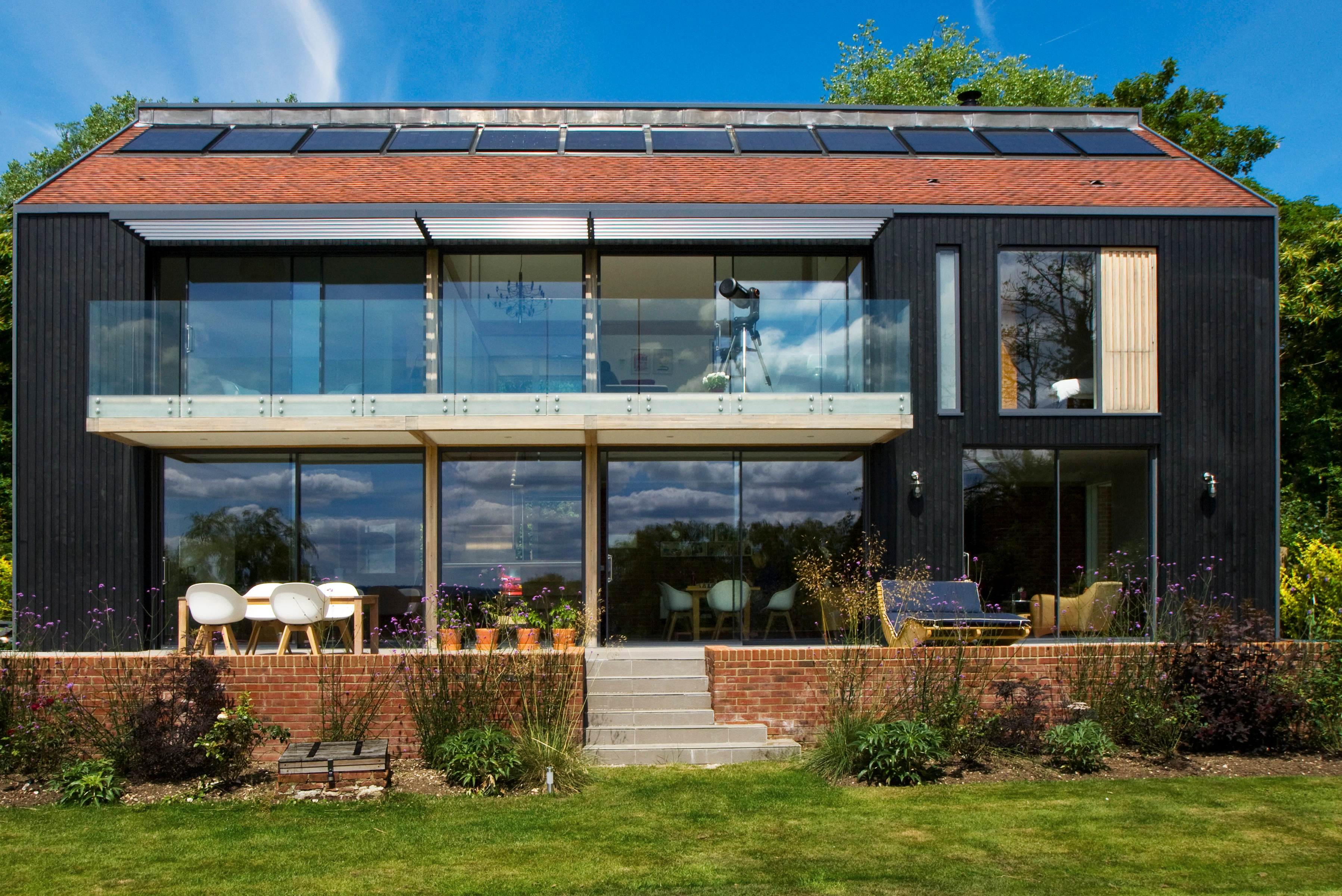 Large glazed windows and doors of the back of a large oak glulam framed house with a raised patio and green lawn in front