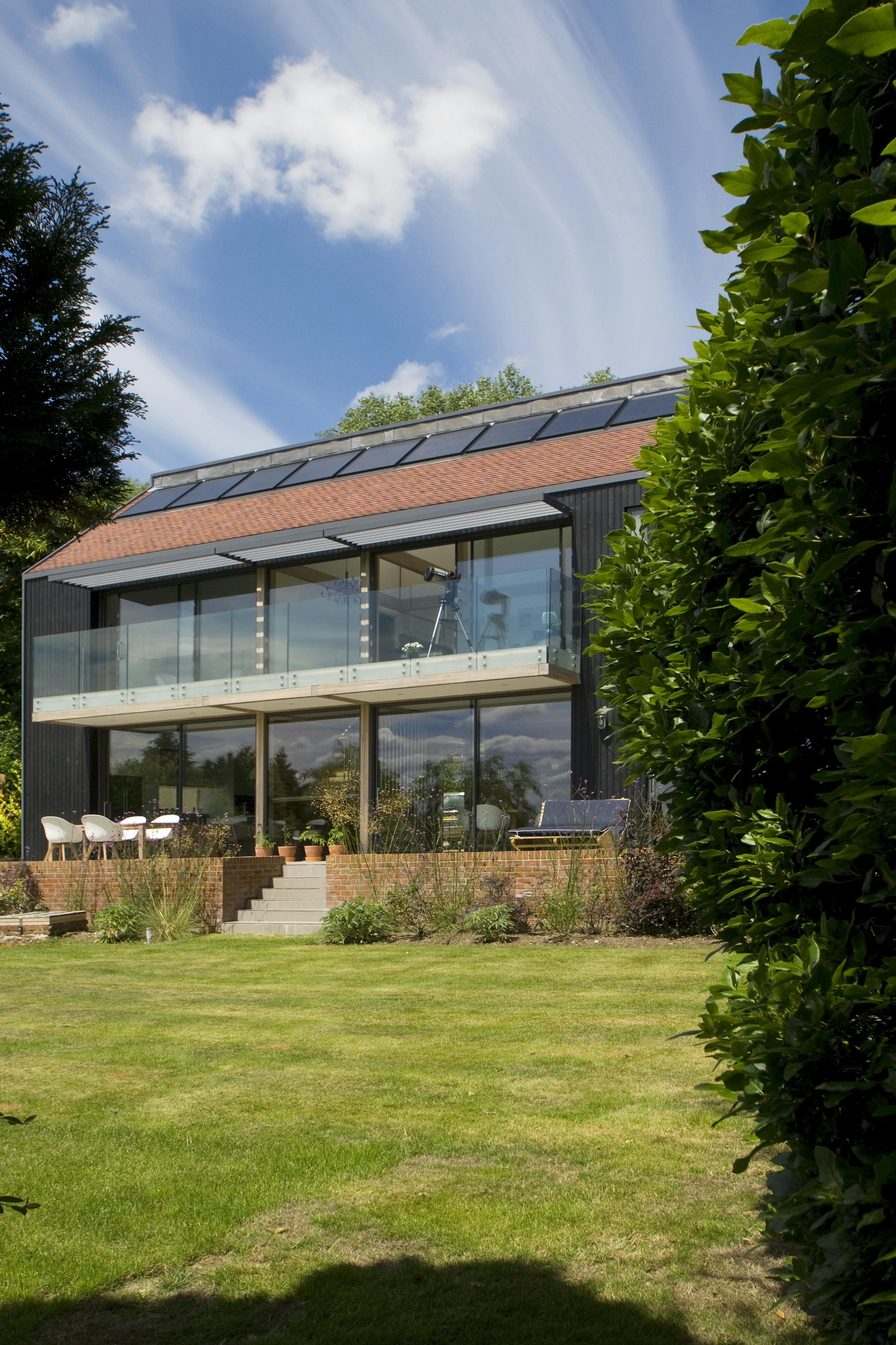 Large glazed windows and doors of the back of a large oak glulam framed house with a raised patio and green lawn in front