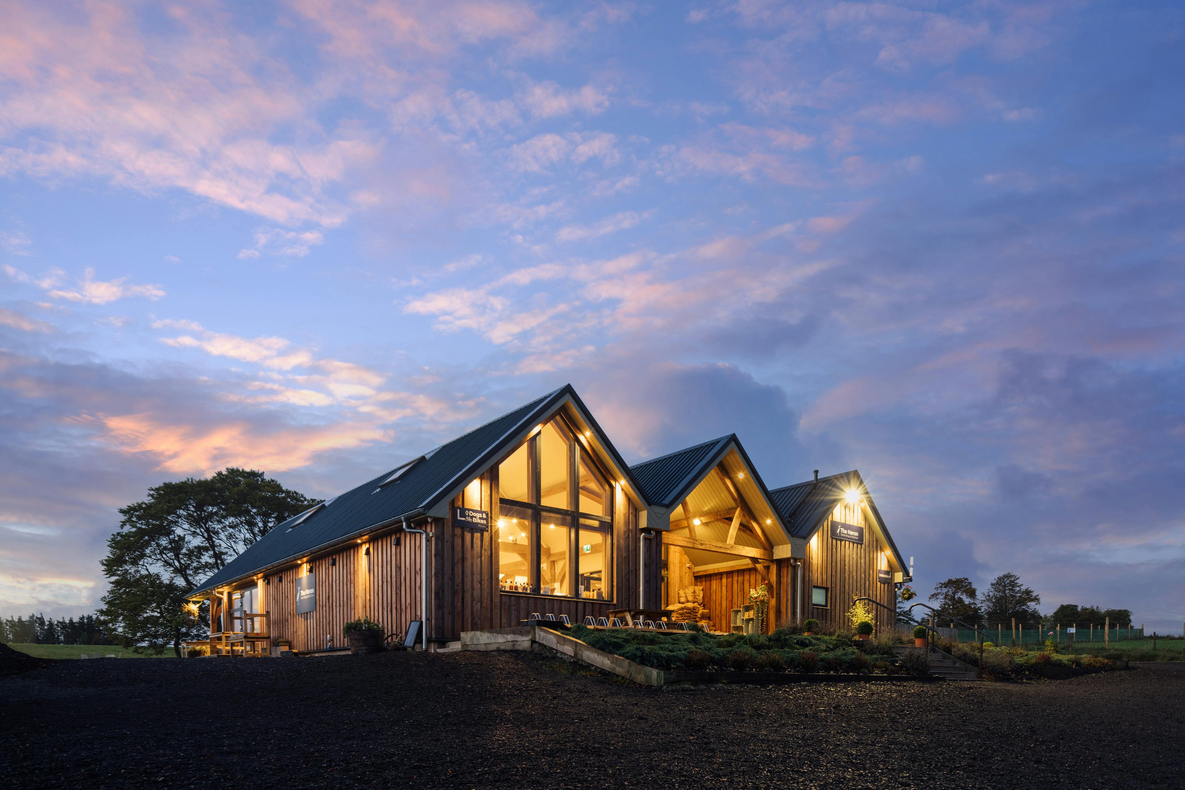 At dusk lights shine from a 3 barn timber clad oak framed farm shop and cafe with an oak framed entrance