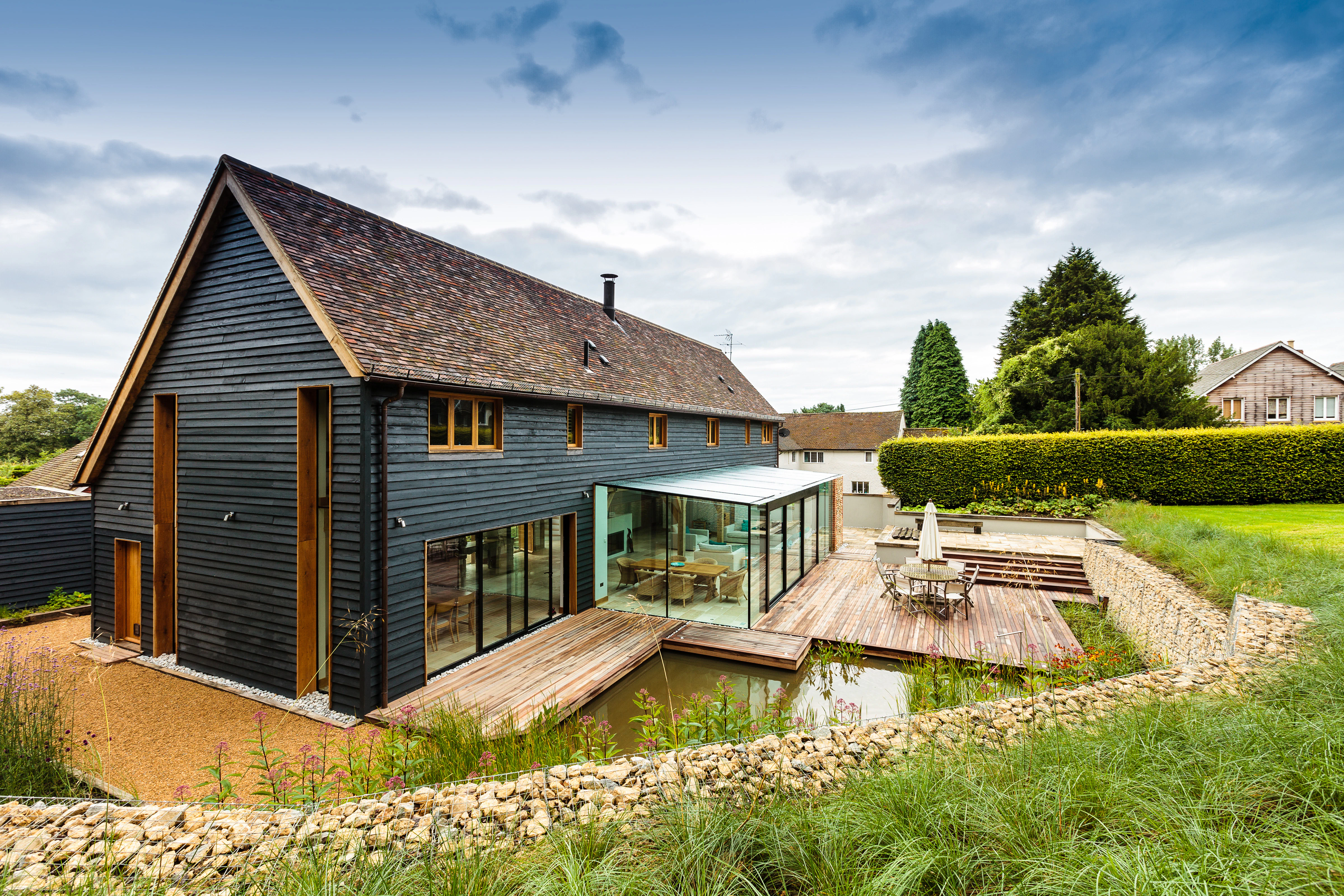 The rear and side of a black clad oak framed house with red roof tiles and a modern glazed extension, with a large landscaped garden, pond and patio