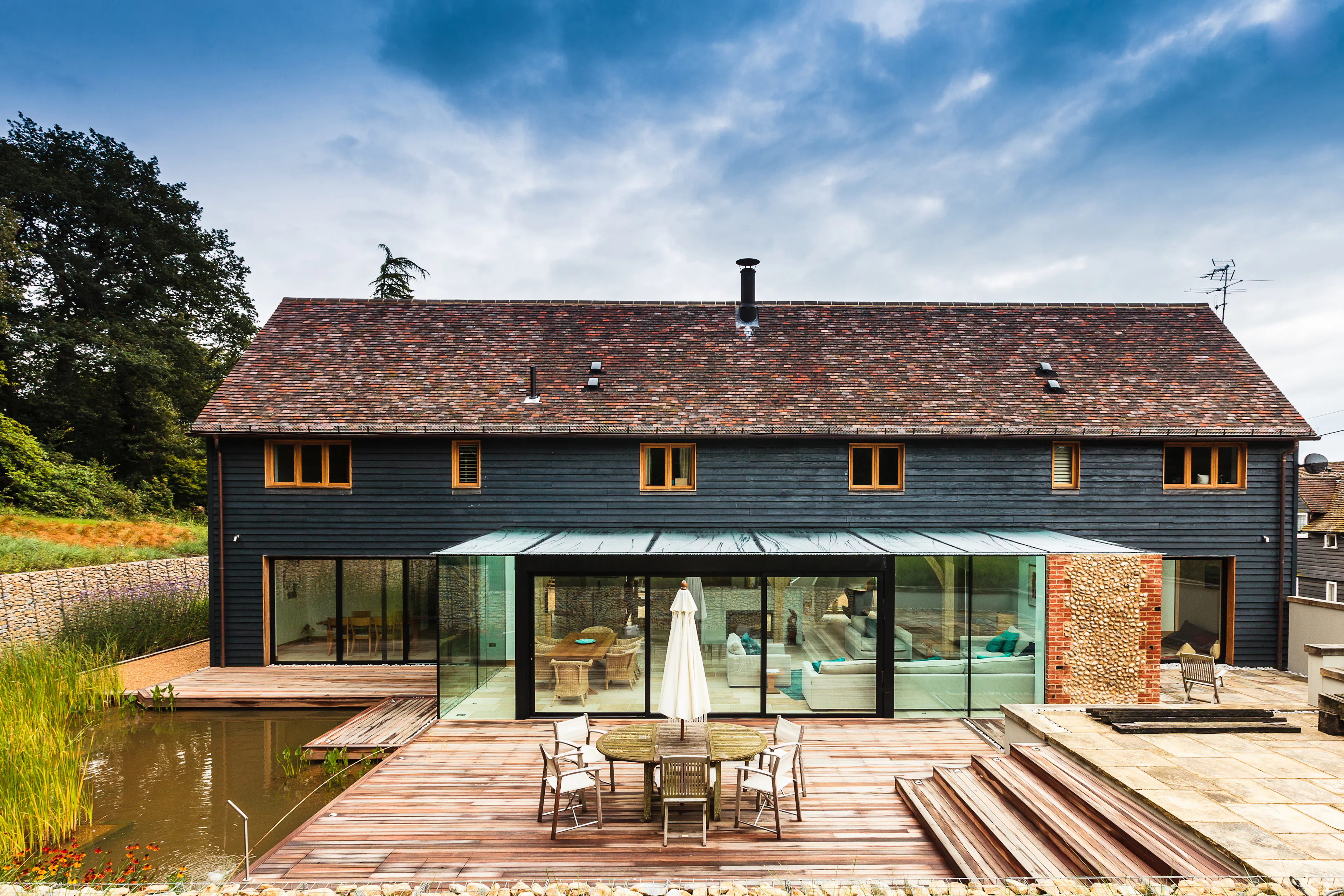 The rear of a black clad oak framed house with red roof tiles and a modern glazed extension, with a large landscaped garden, pond and patio