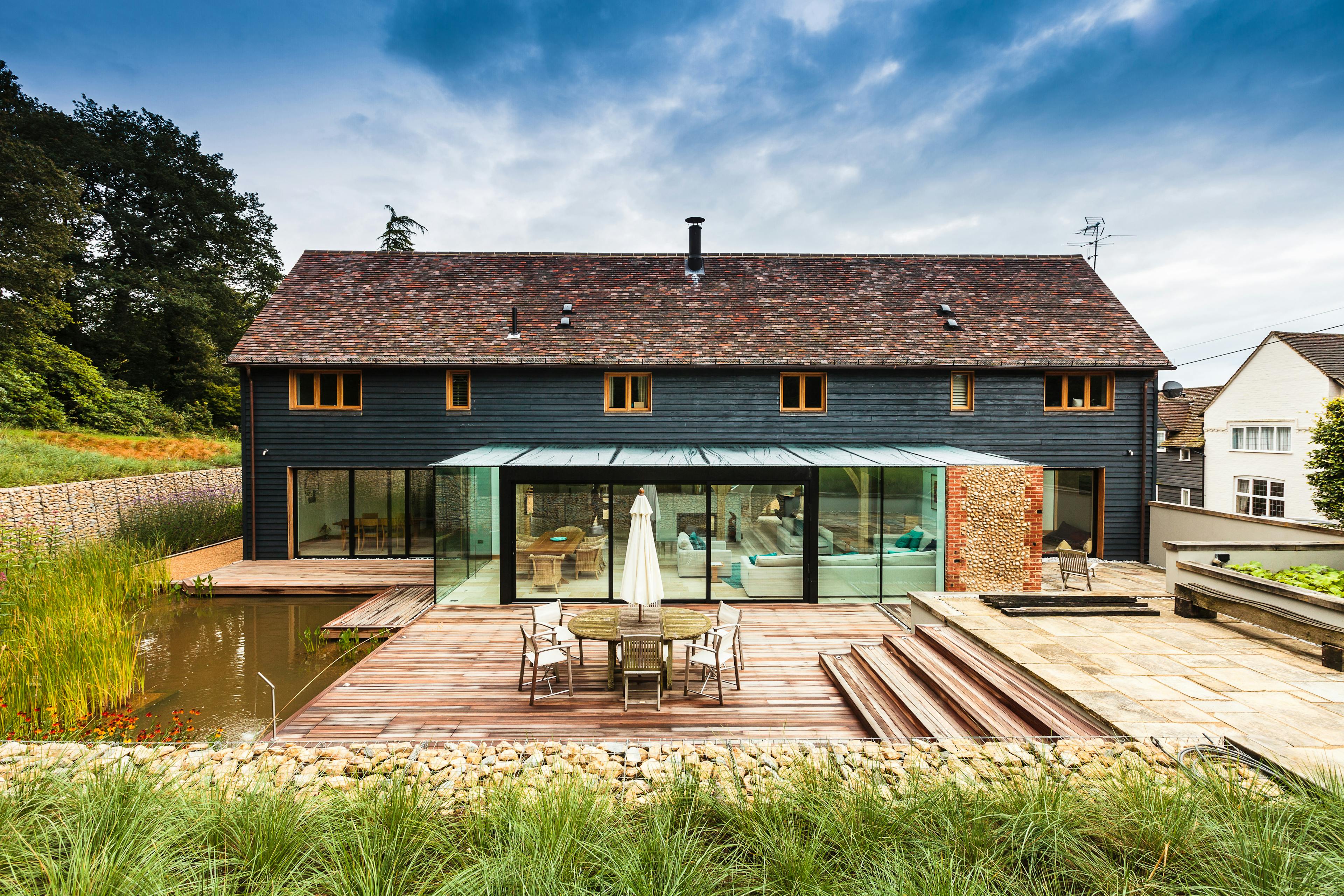 The rear of a black clad oak framed house with red roof tiles and a modern glazed extension, with a large landscaped garden, pond and patio