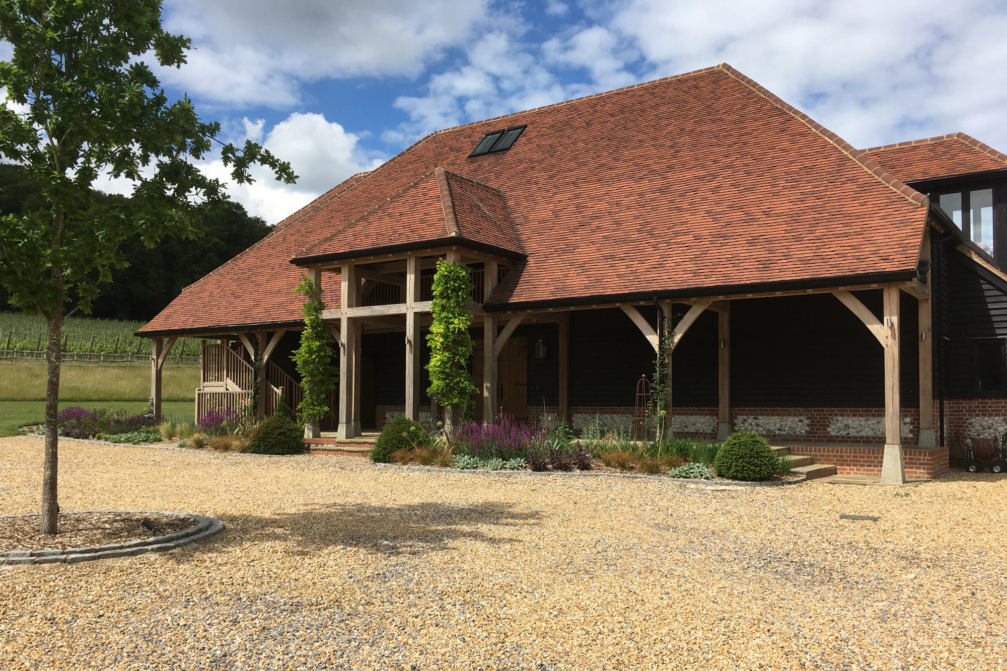 A large winery building with red roof tiles and oak framed balconies and veranda nestled in a green valley and a gravel driveway in front