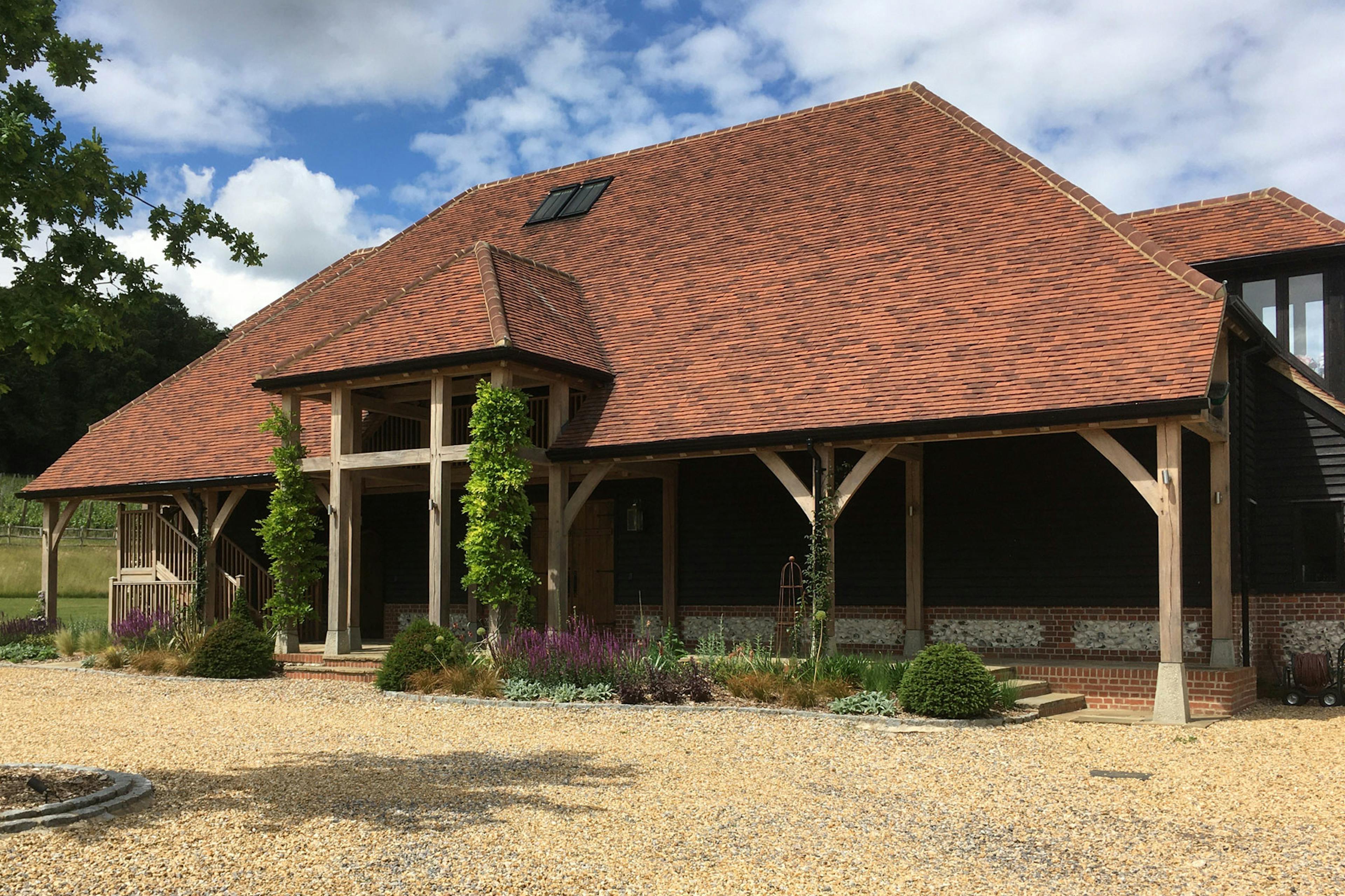 A large winery building with red roof tiles and oak framed balconies and veranda nestled in a green valley and a gravel driveway in front