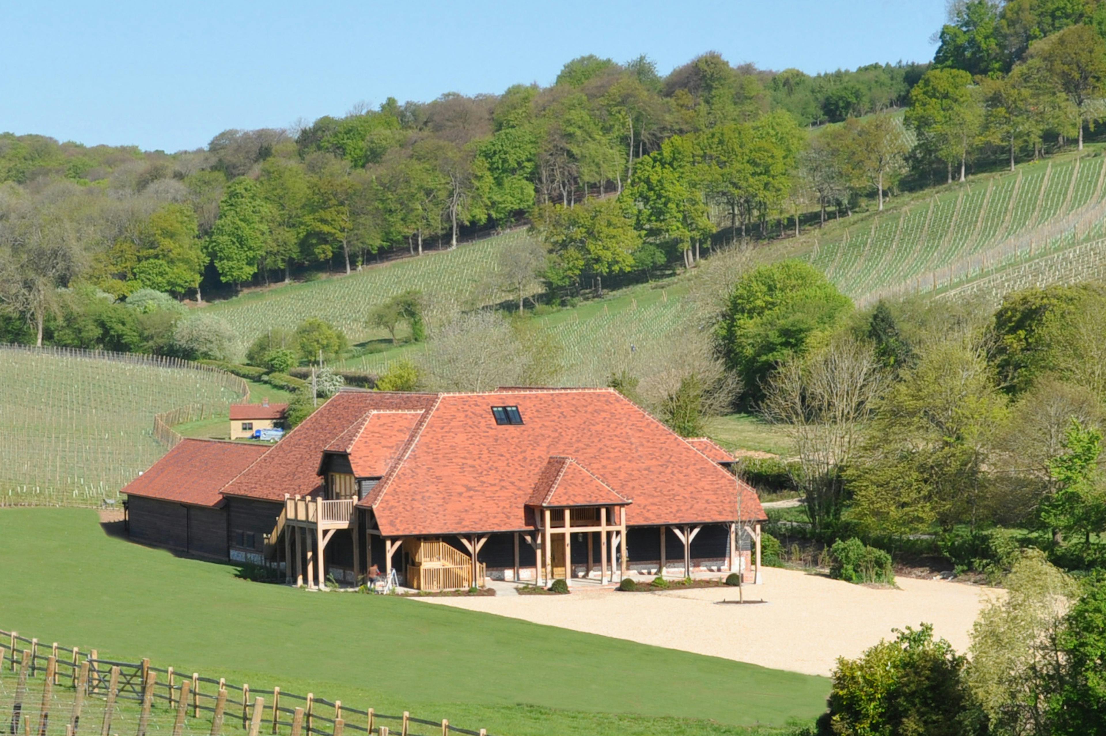 A large winery building with red roof tiles and oak framed balconies and veranda nestled in a green valley