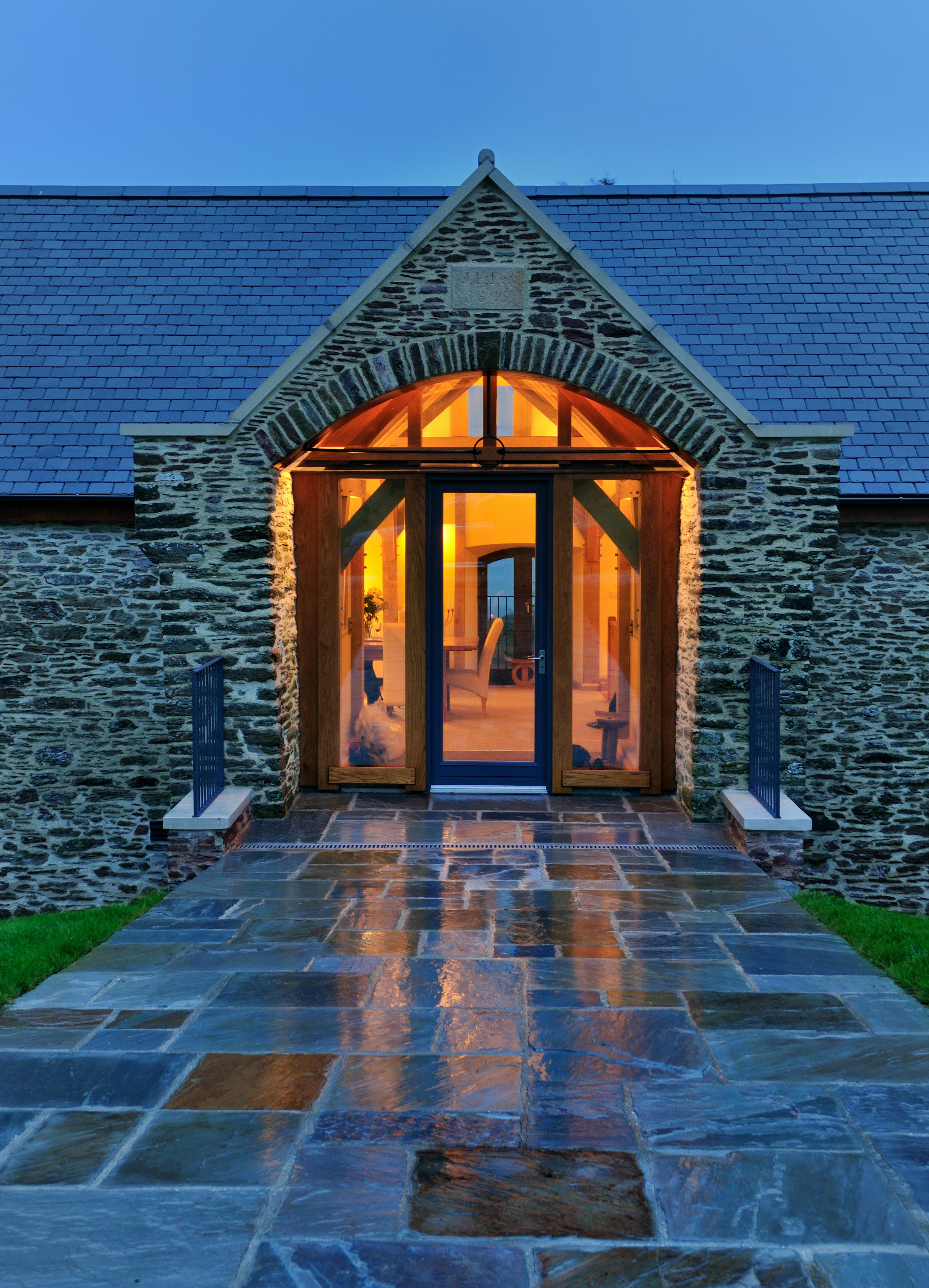 At dusk light shines from the glazed entrance of a stone clad barn converted to a home