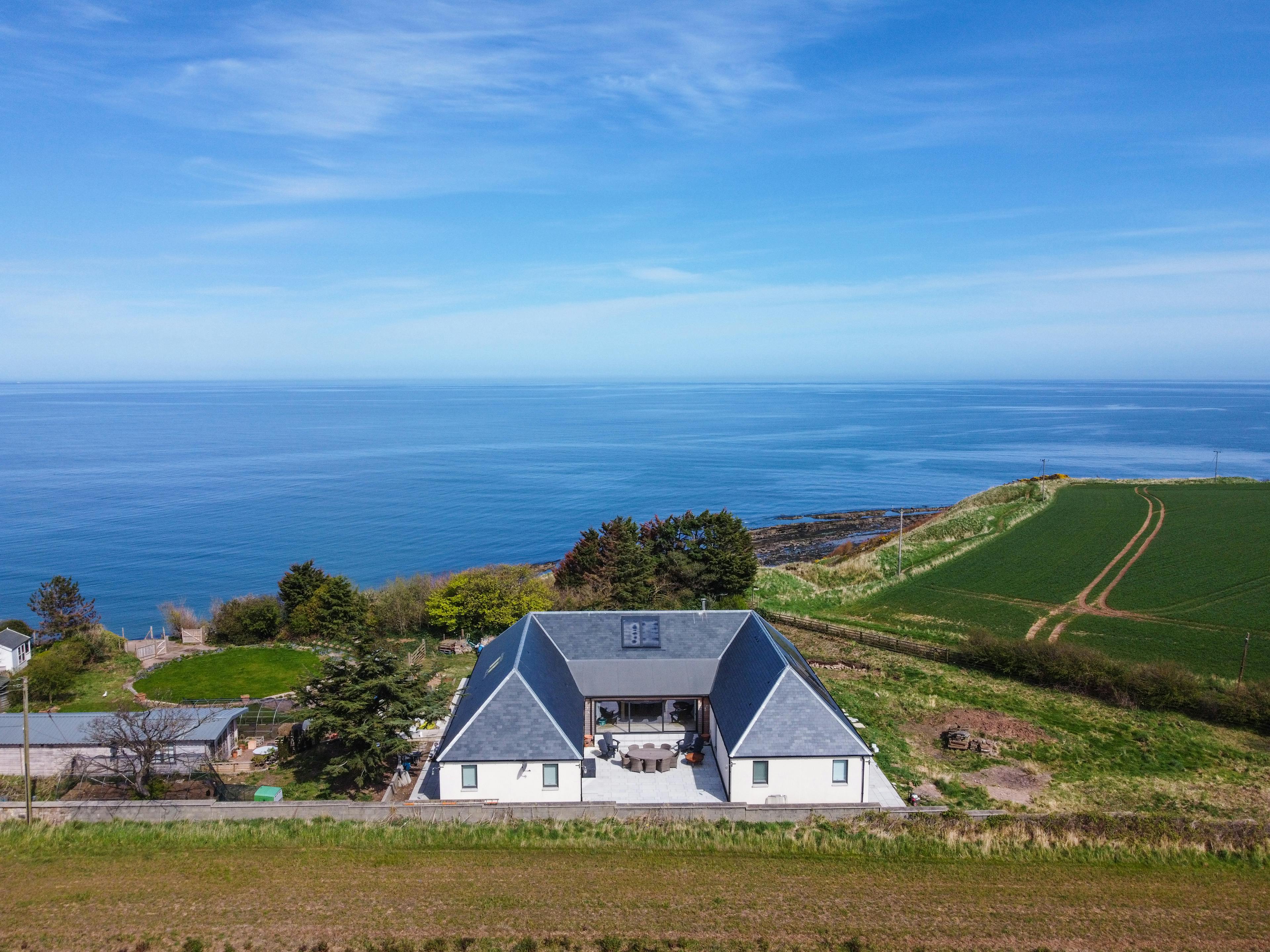 A single storey u-shaped home with a black slate sloping roof near the edge of a cliff, surrounded with green fields and with the blue sea in the background