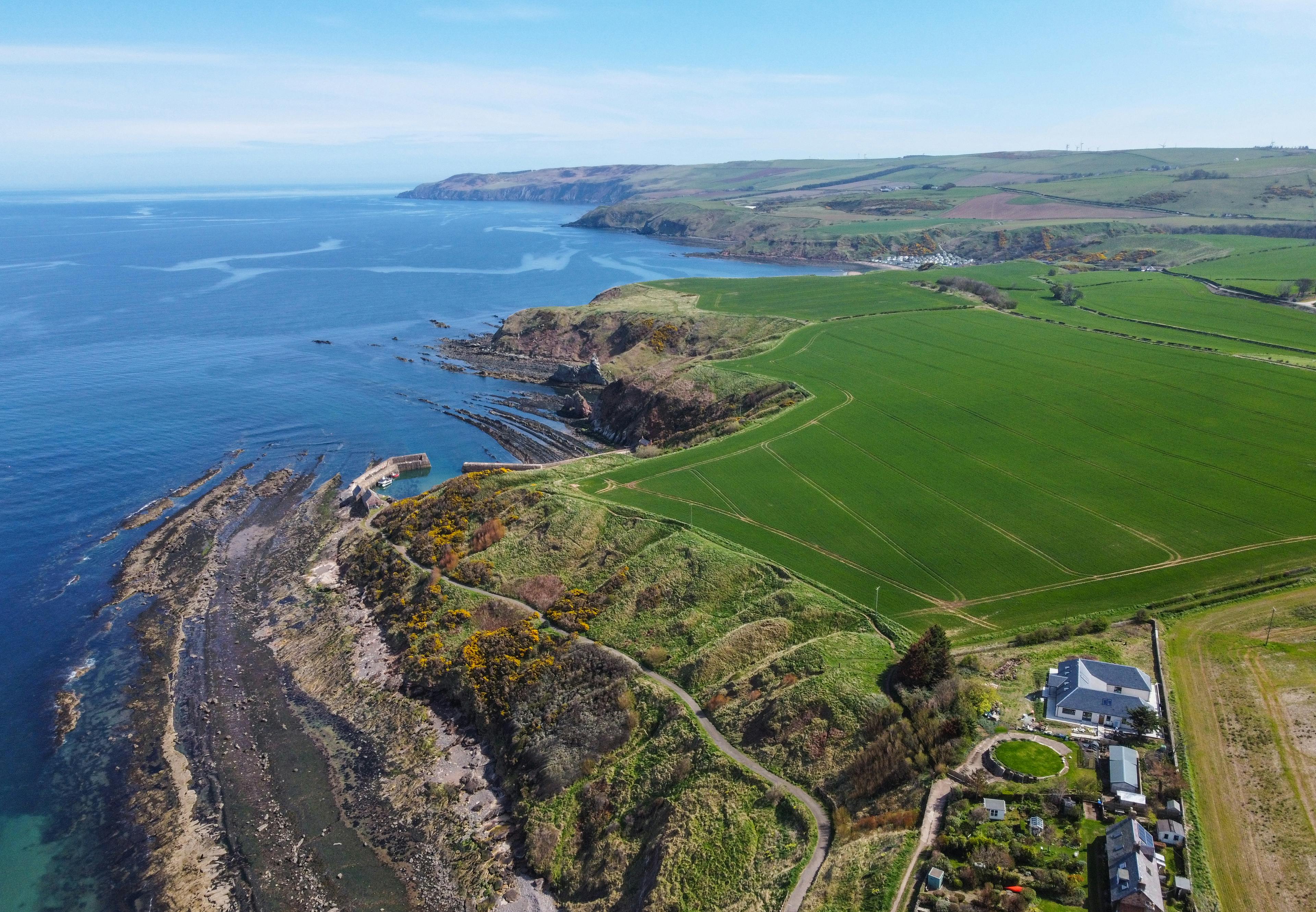 A single storey u-shaped home with a black slate sloping roof near the edge of a cliff, surrounded with green fields and with the blue sea in the background