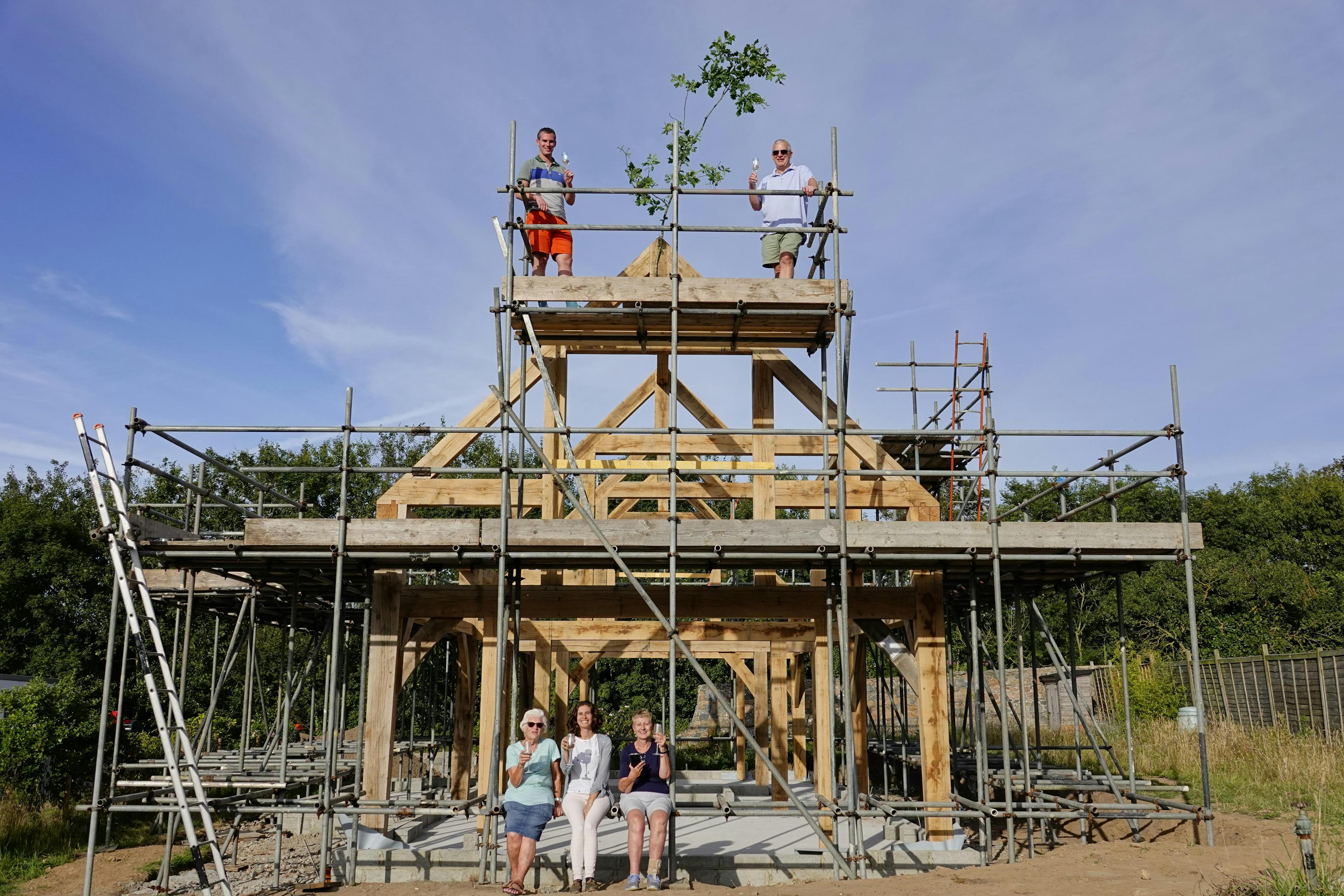 Some carpenters on scaffolding at the top of an oak frame during installation on a construction site with blue skies behind