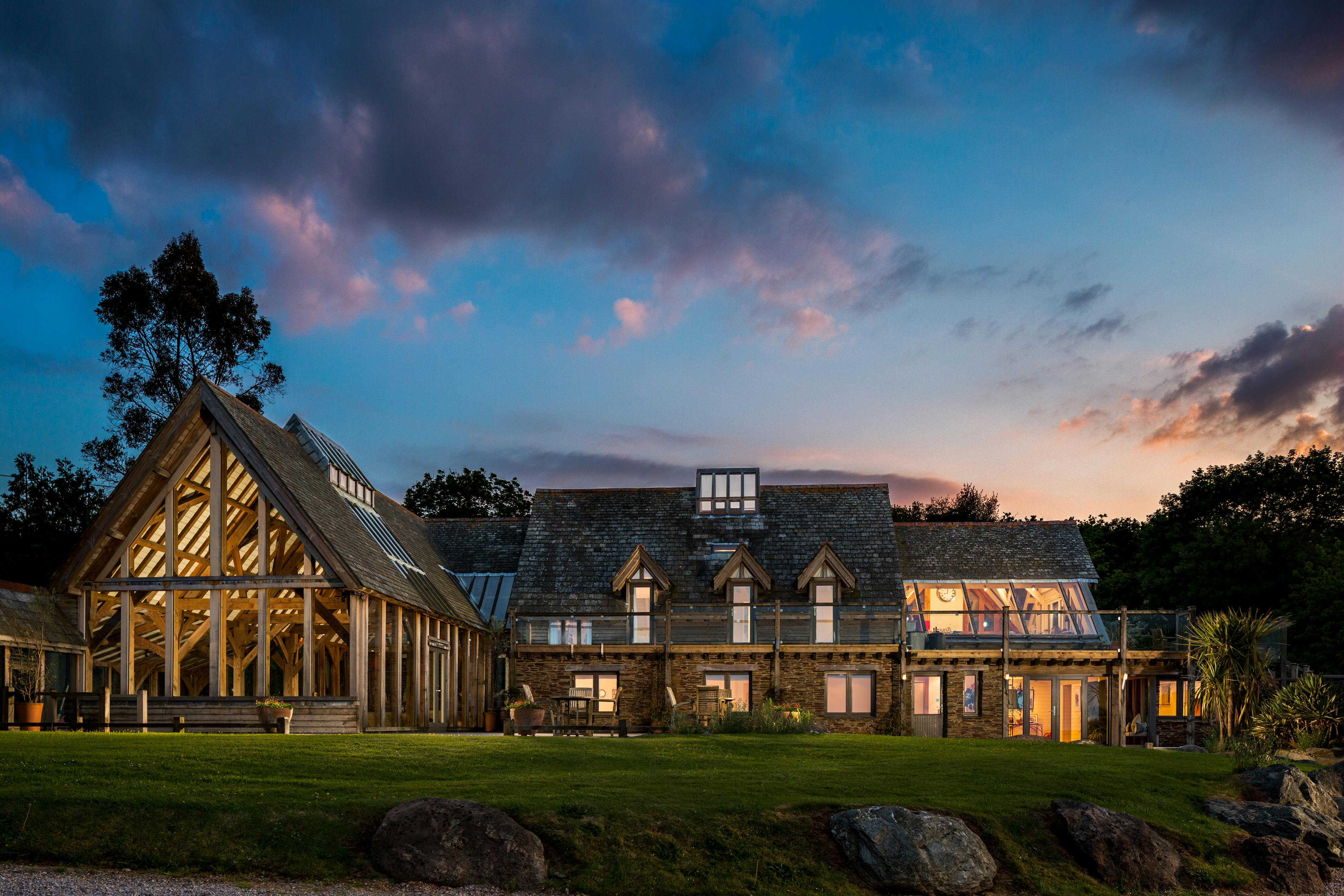 At dusk lights shine from the windows of a large oak framed building with a lawn