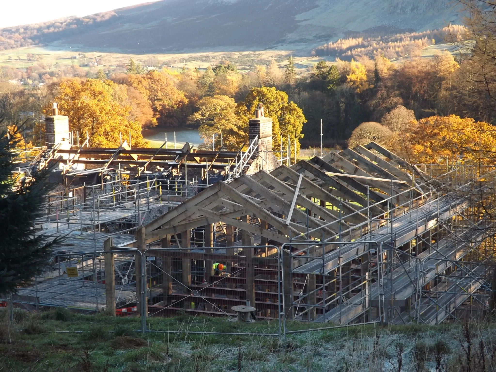 An oak frame with scissor trusses and a red steel frame on a construction site during installation, nestled in a green valley