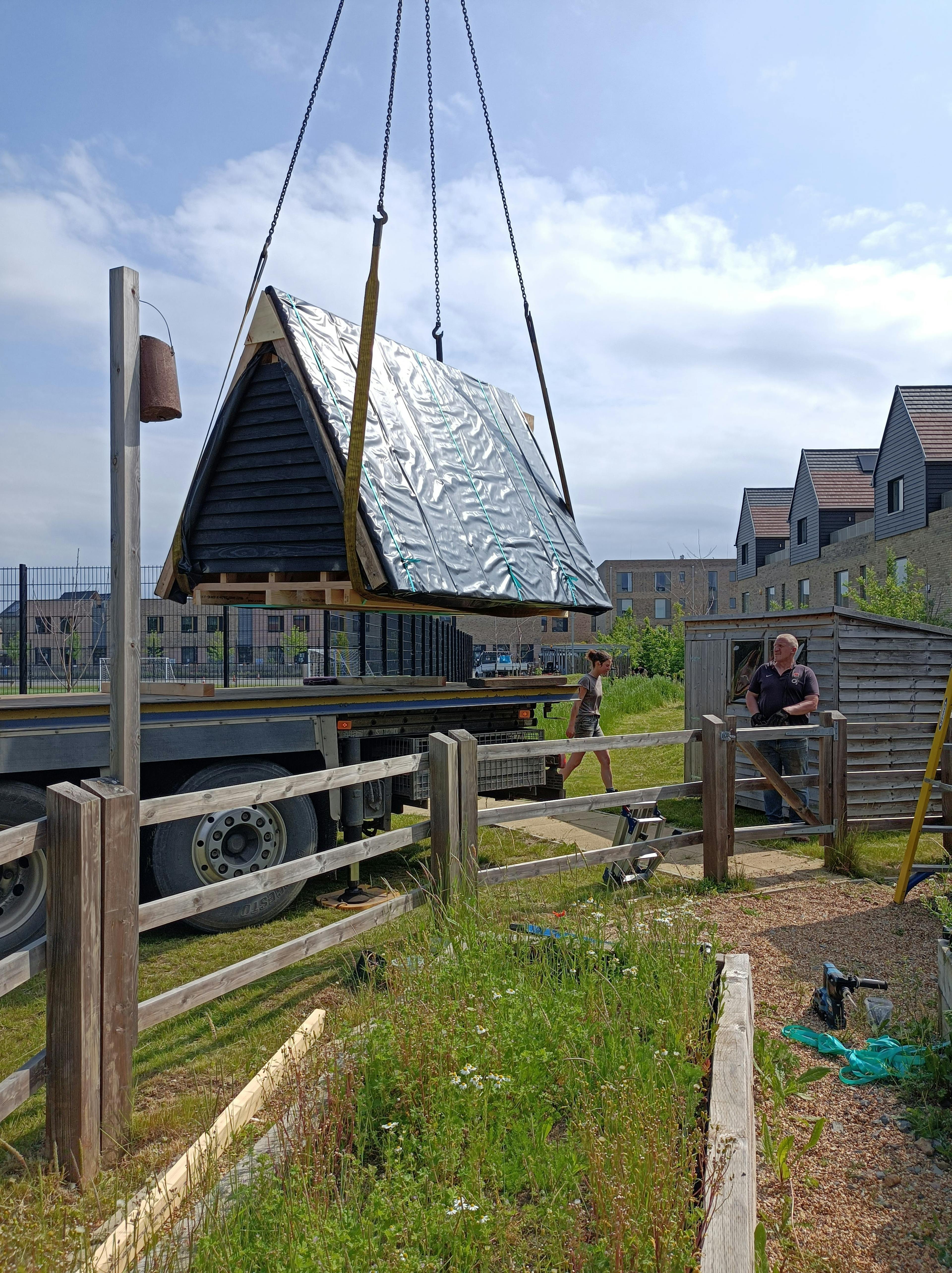 A crane lowers the roof of a black clad oak framed cabin with reflective steel roof