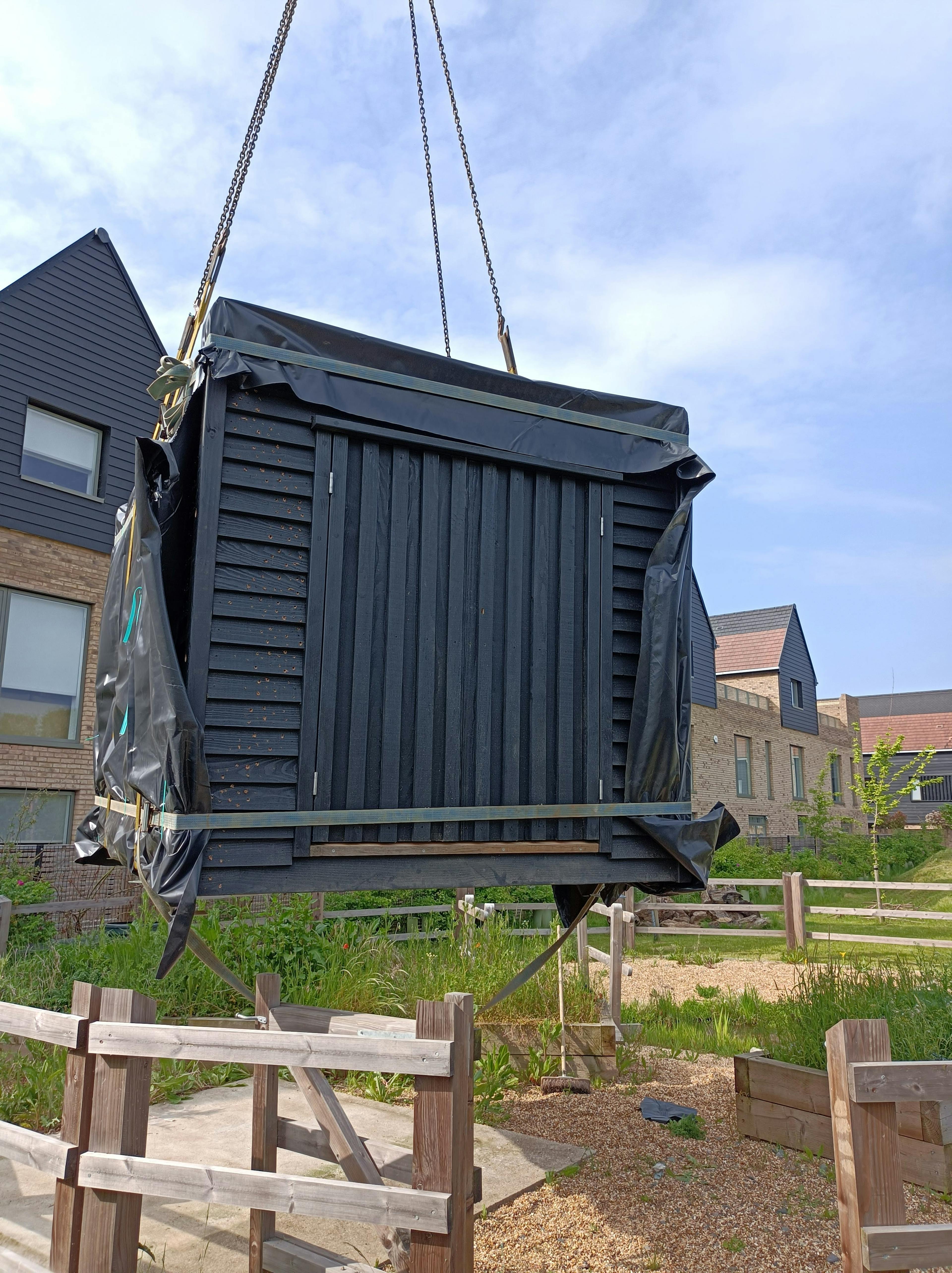 A crane lowers the bottom half of a black clad oak framed cabin with reflective steel roof