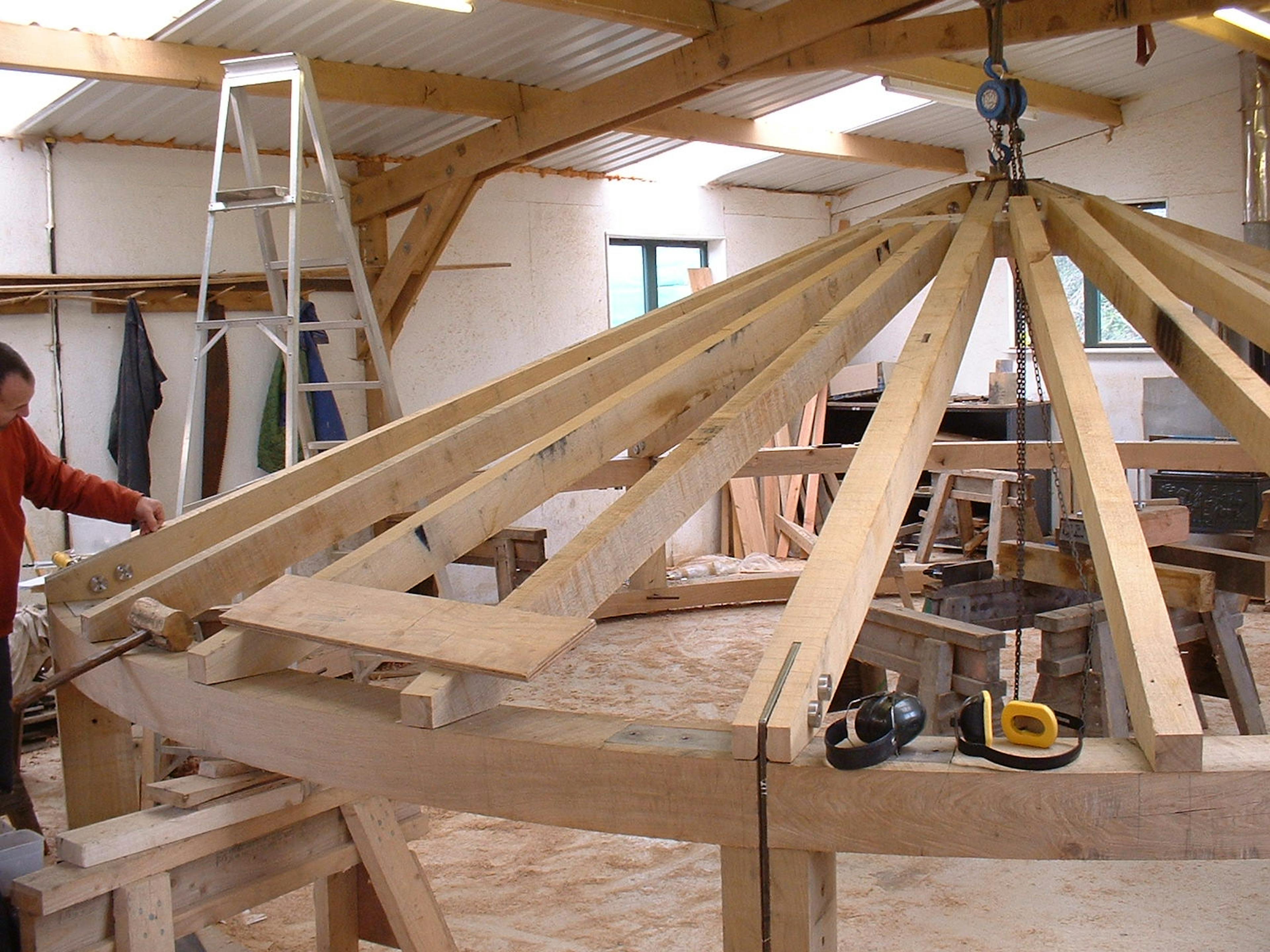 Timber roof structure under construction inside a workshop, with visible beams and joints being assembled by craftsmen