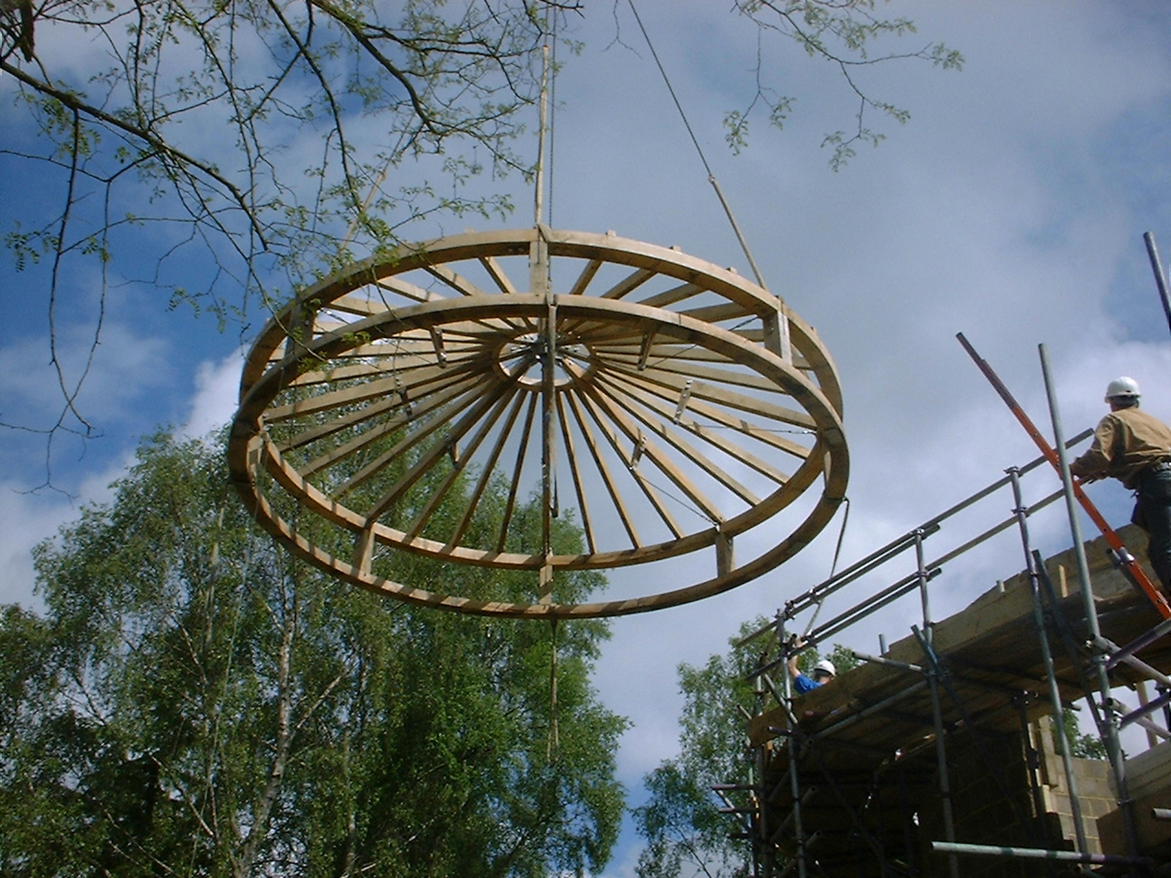 Construction site image showing a crane lifting a cylindrical oak timber component into place as part of the roof structure assembly of an oak-framed house