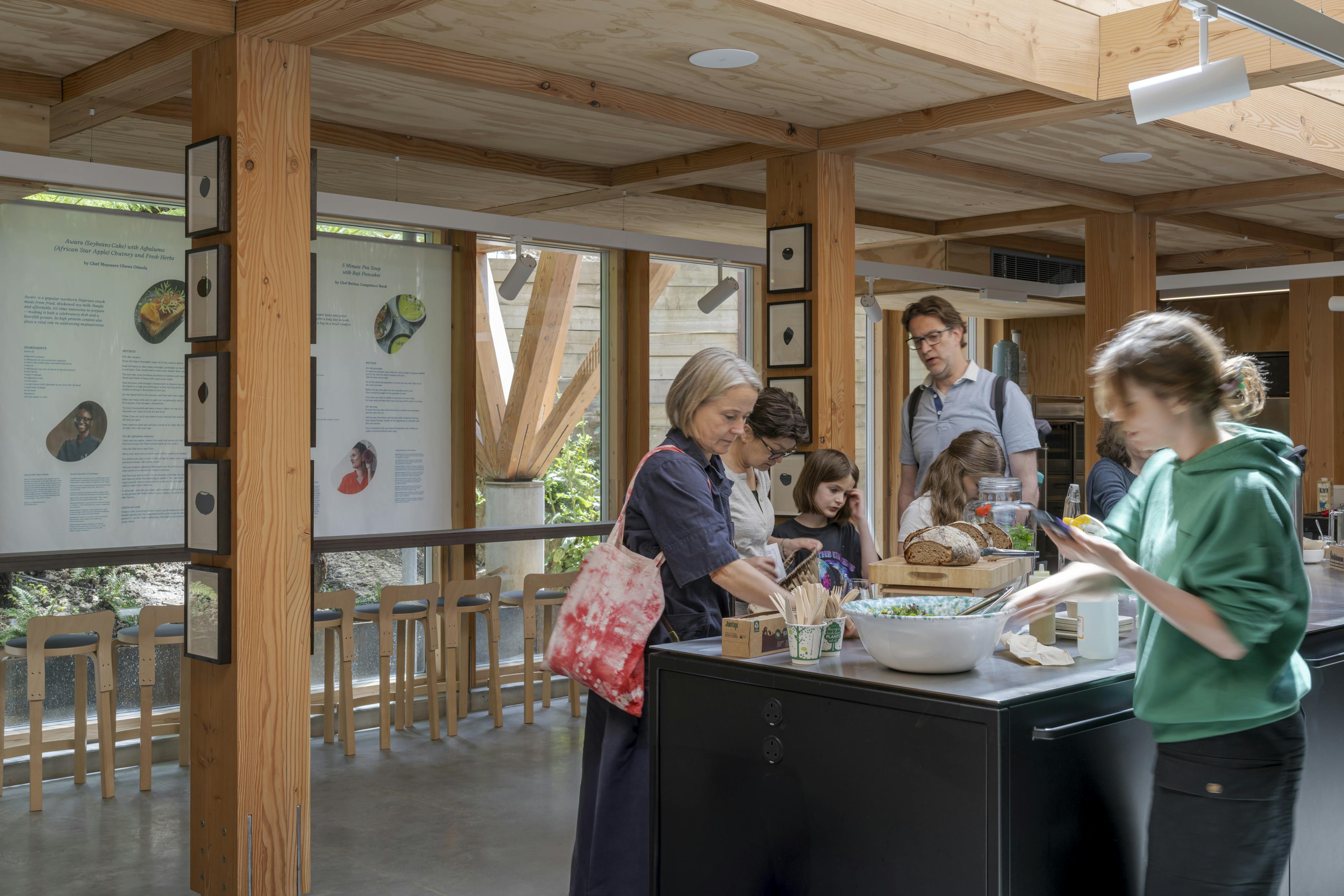 Interior of a Douglas fir-framed event space featuring a vaulted ceiling with glazing, set up for a seminar and exhibition, including a visible kitchen area