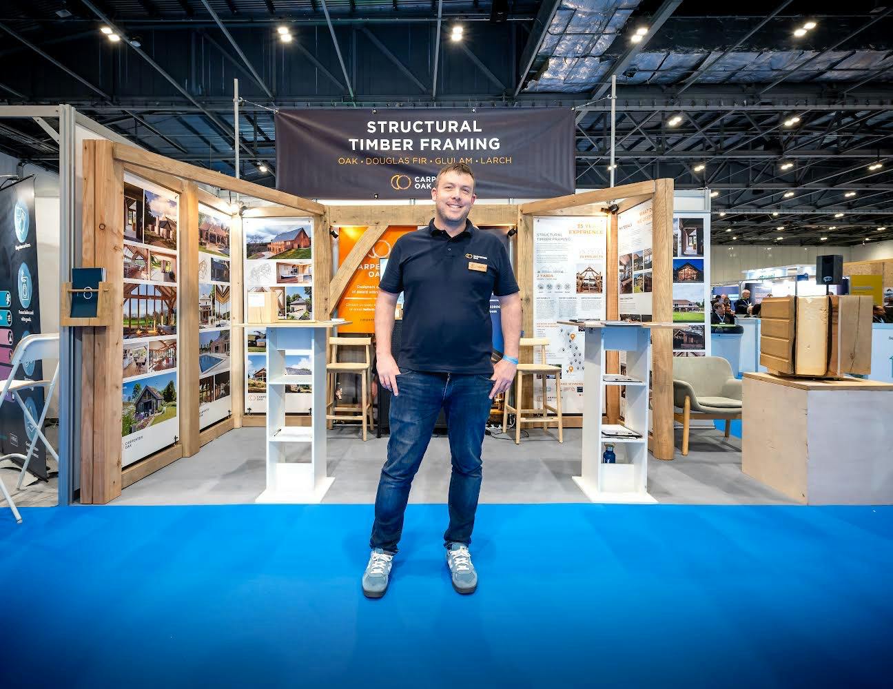 A sales person in front of a timber framed show stand at a self build show