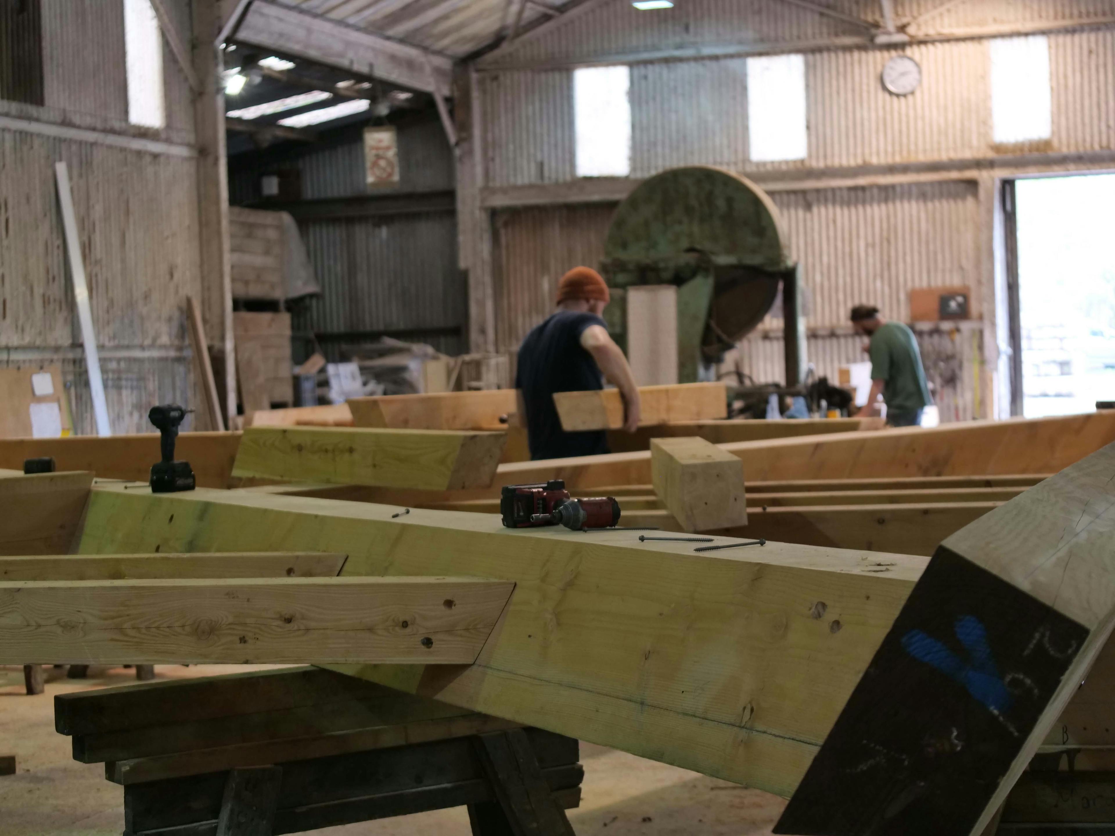 Two carpenters work on a frame for a boathouse in a large workshop