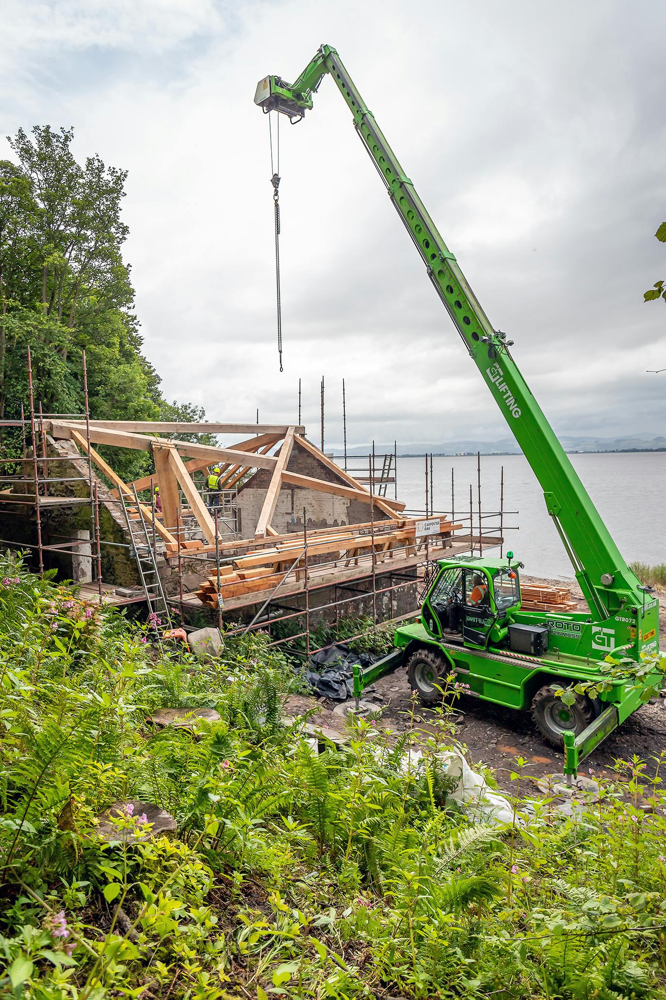 A frame being installed in a boathouse on the shore of a river - a crane lifts a piece of timber into place
