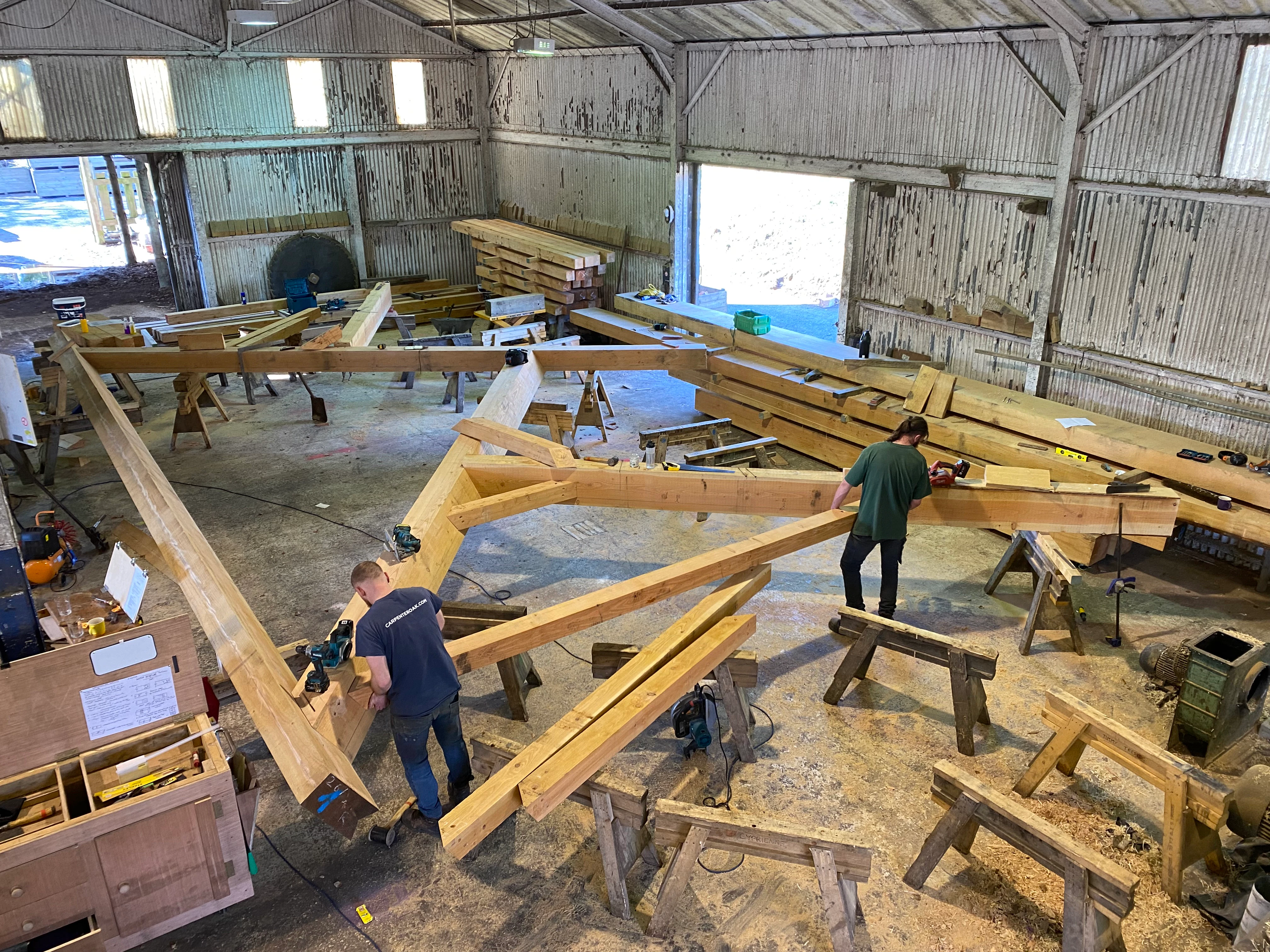 Two carpenters work on a large timber frame for a boathouse in a large workshop