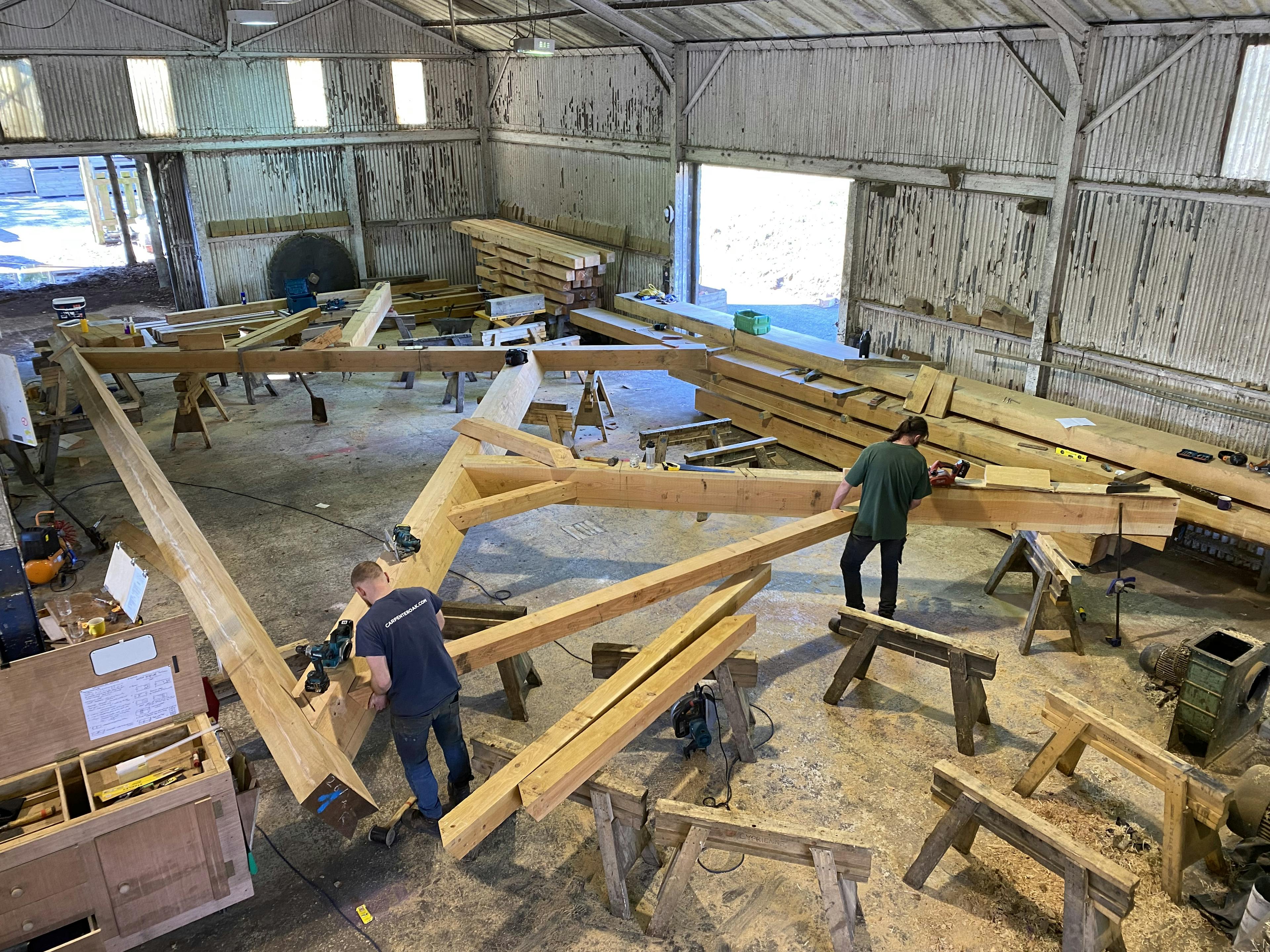 Two carpenters work on a large timber frame for a boathouse in a large workshop
