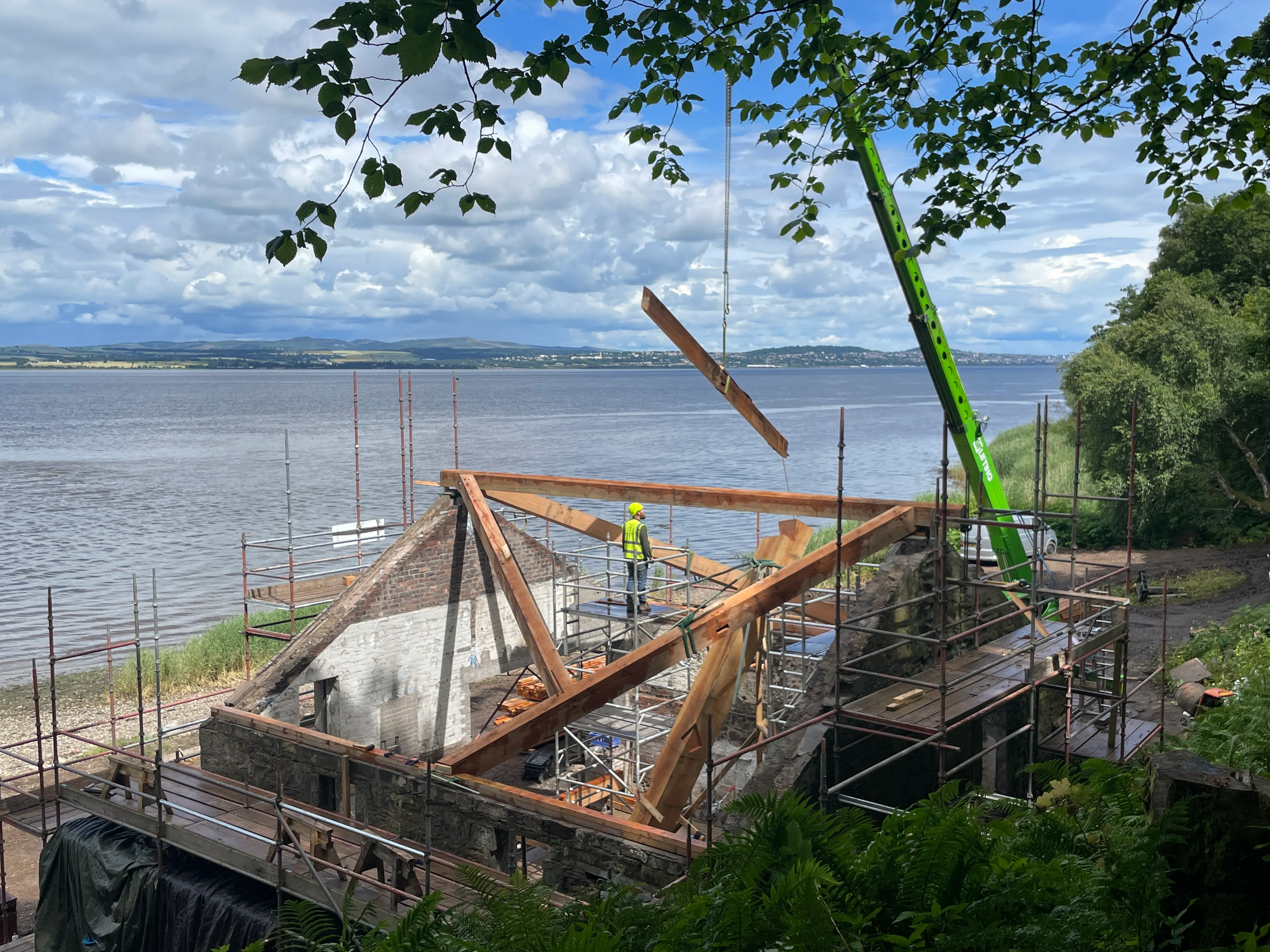 A frame being installed in a boathouse on the shore of a river - a crane lifts a piece of timber into place