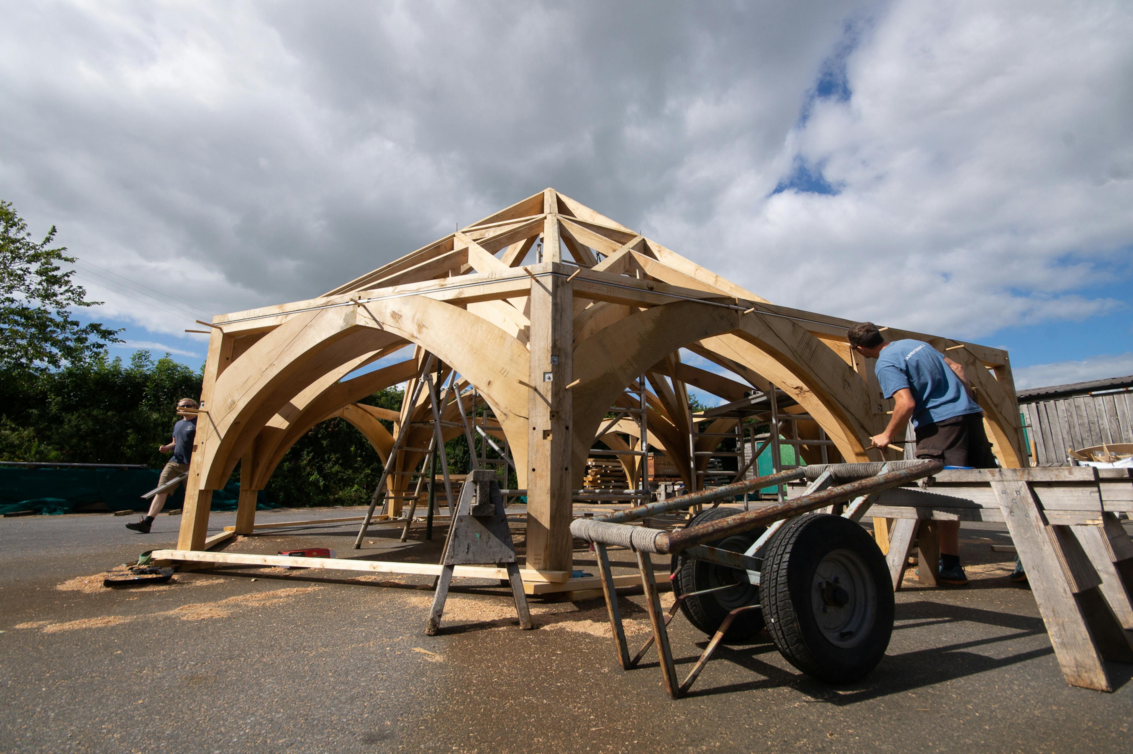 Timber frame for Radley College under construction in the Carpenter Oak workshop, designed by Purcell Architects, with craftspeople assembling large wooden beams and joints.