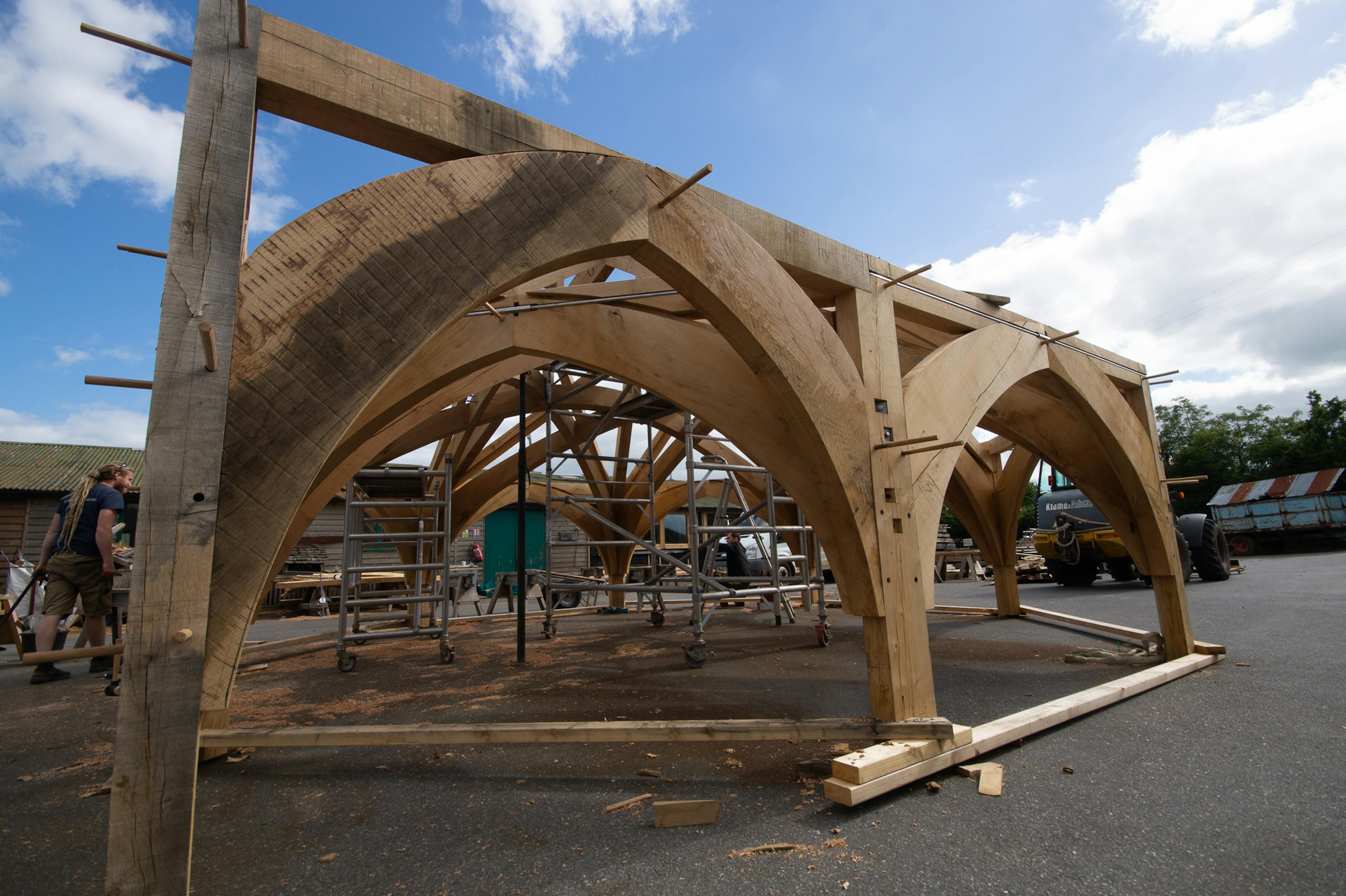 Low-angle view of the timber frame for Radley College under construction in the Carpenter Oak workshop, designed by Purcell Architects, emphasizing the scale and structure of the wooden beams.