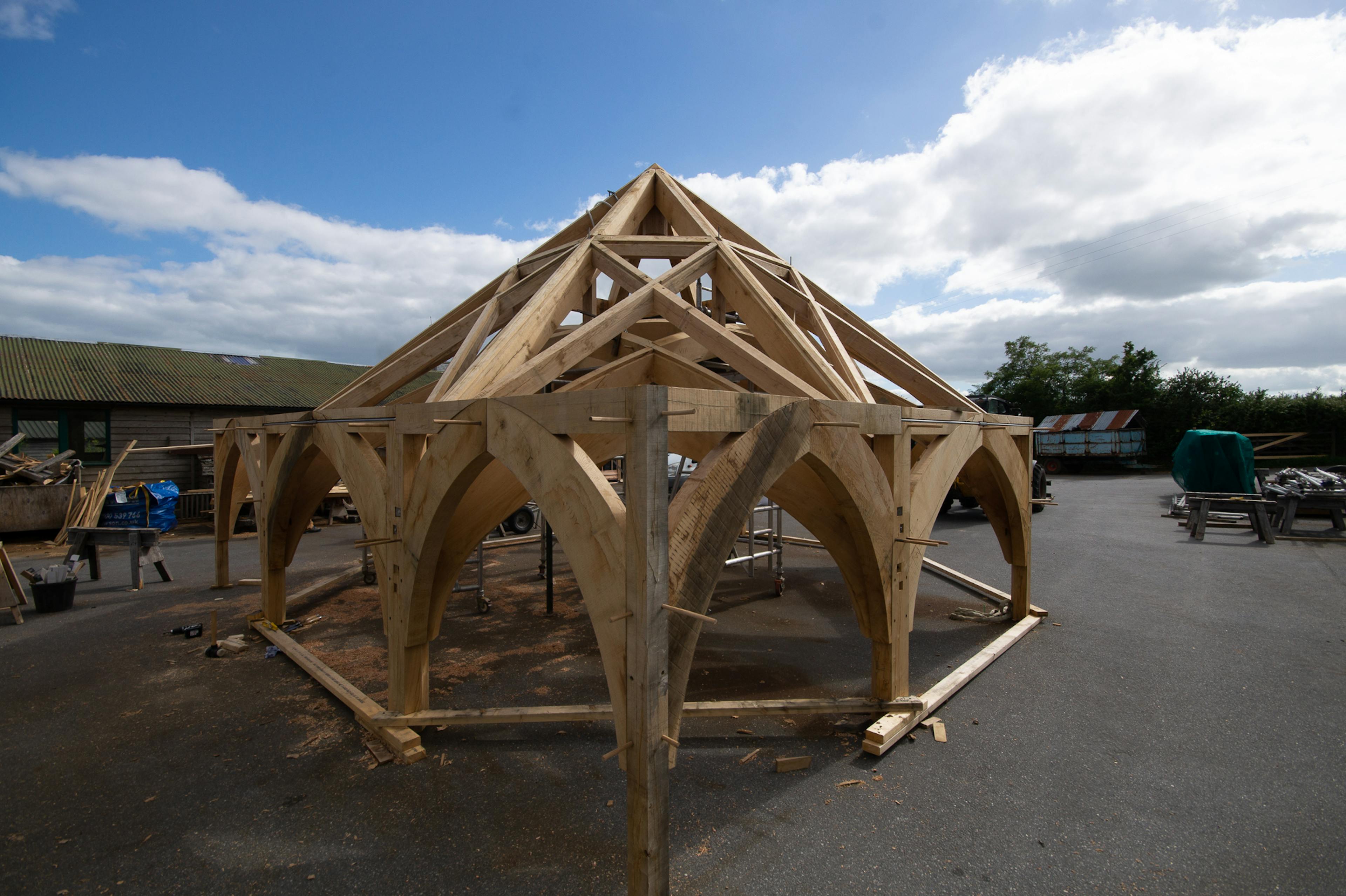 Upward view of the timber frame for Radley College under construction in the Carpenter Oak workshop, designed by Purcell Architects, highlighting the geometry and joinery of the wooden beams.