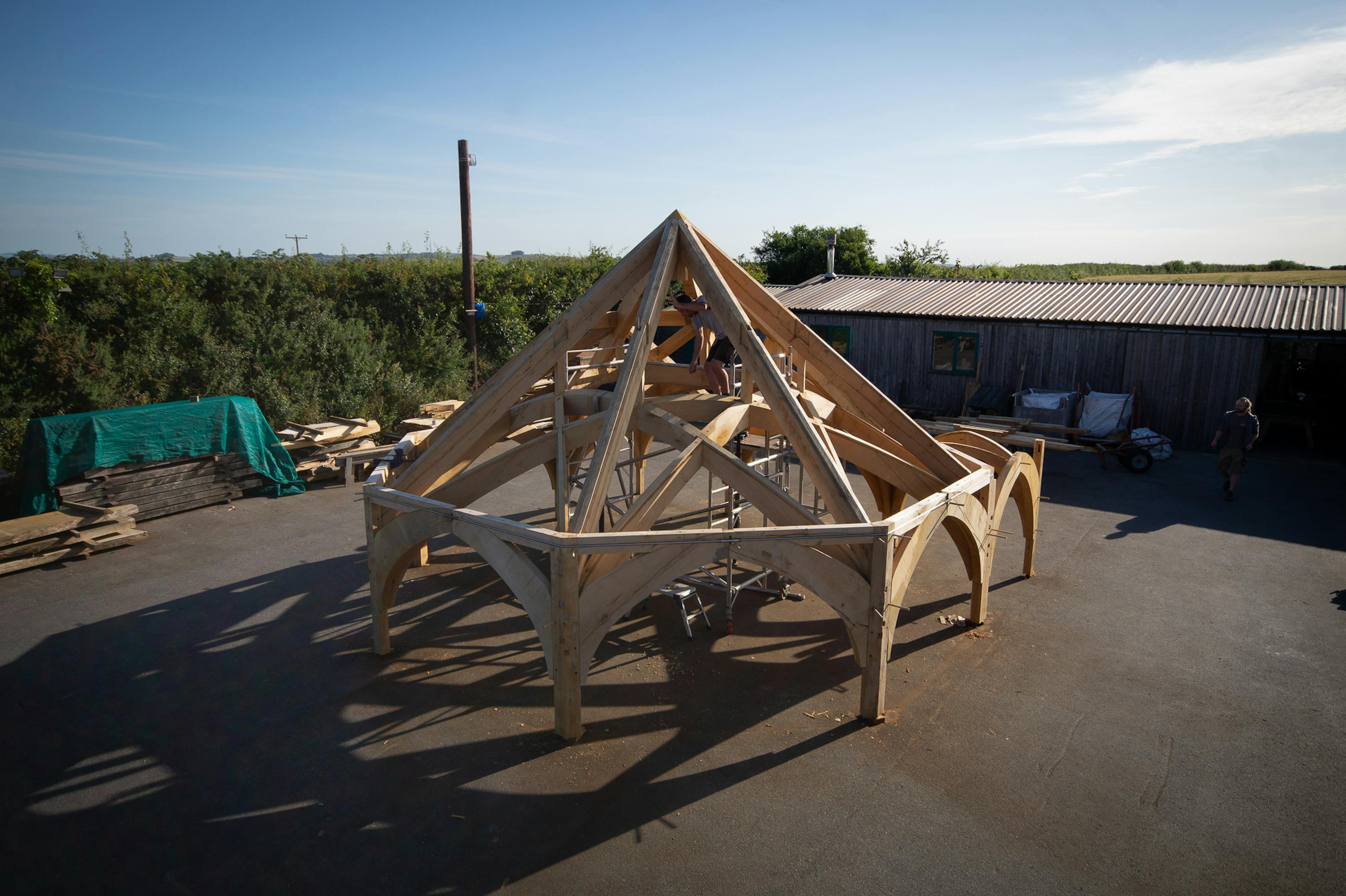 Timber frame for Radley College being assembled in the Carpenter Oak workshop, designed by Purcell Architects, showing workers fitting large wooden beams and joints.