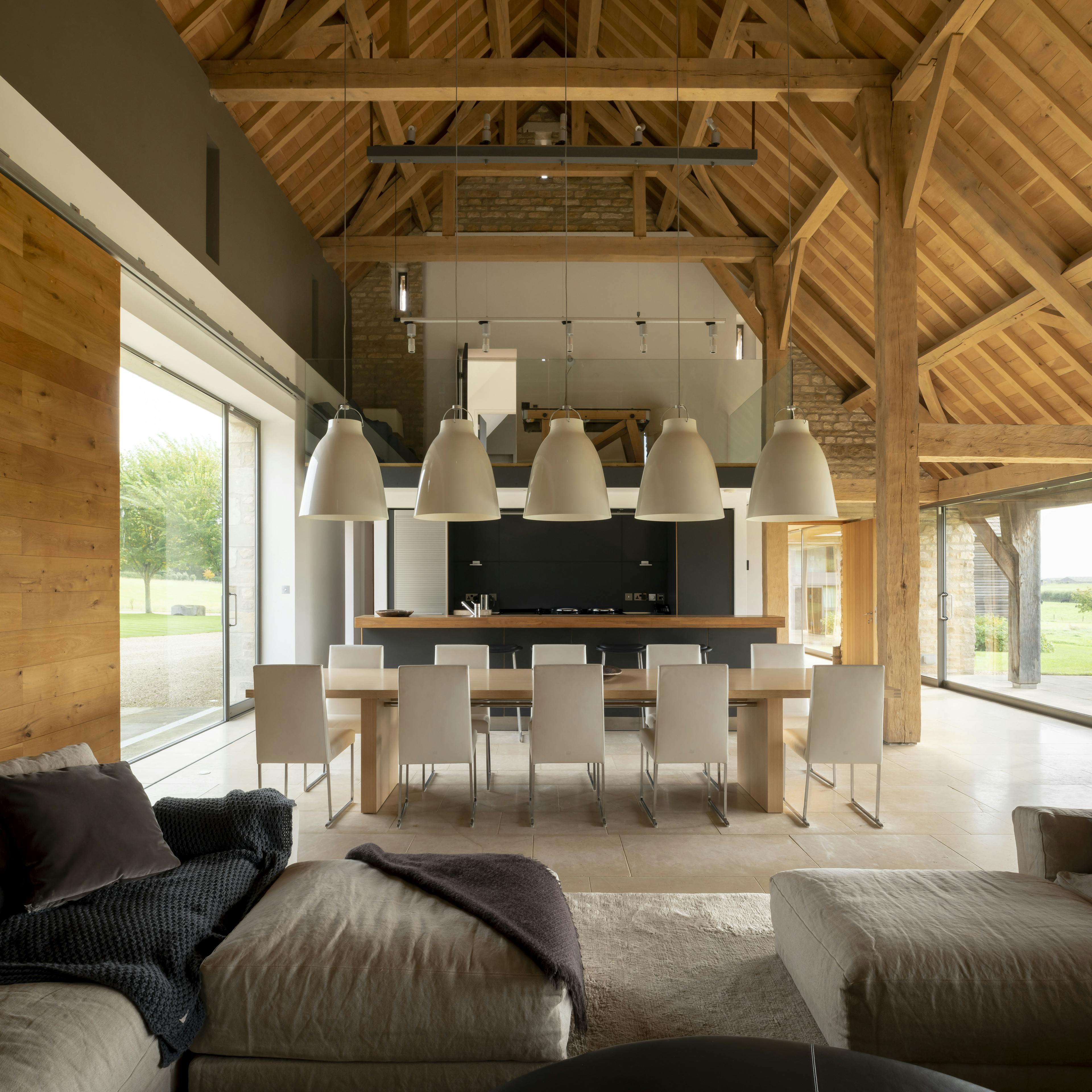 Interior of a renovated oak-framed barn with vaulted ceilings and exposed beams, featuring a contemporary open-plan living area where modern pendant lights hang over the dining space, creating a striking contrast with the traditional oak structure