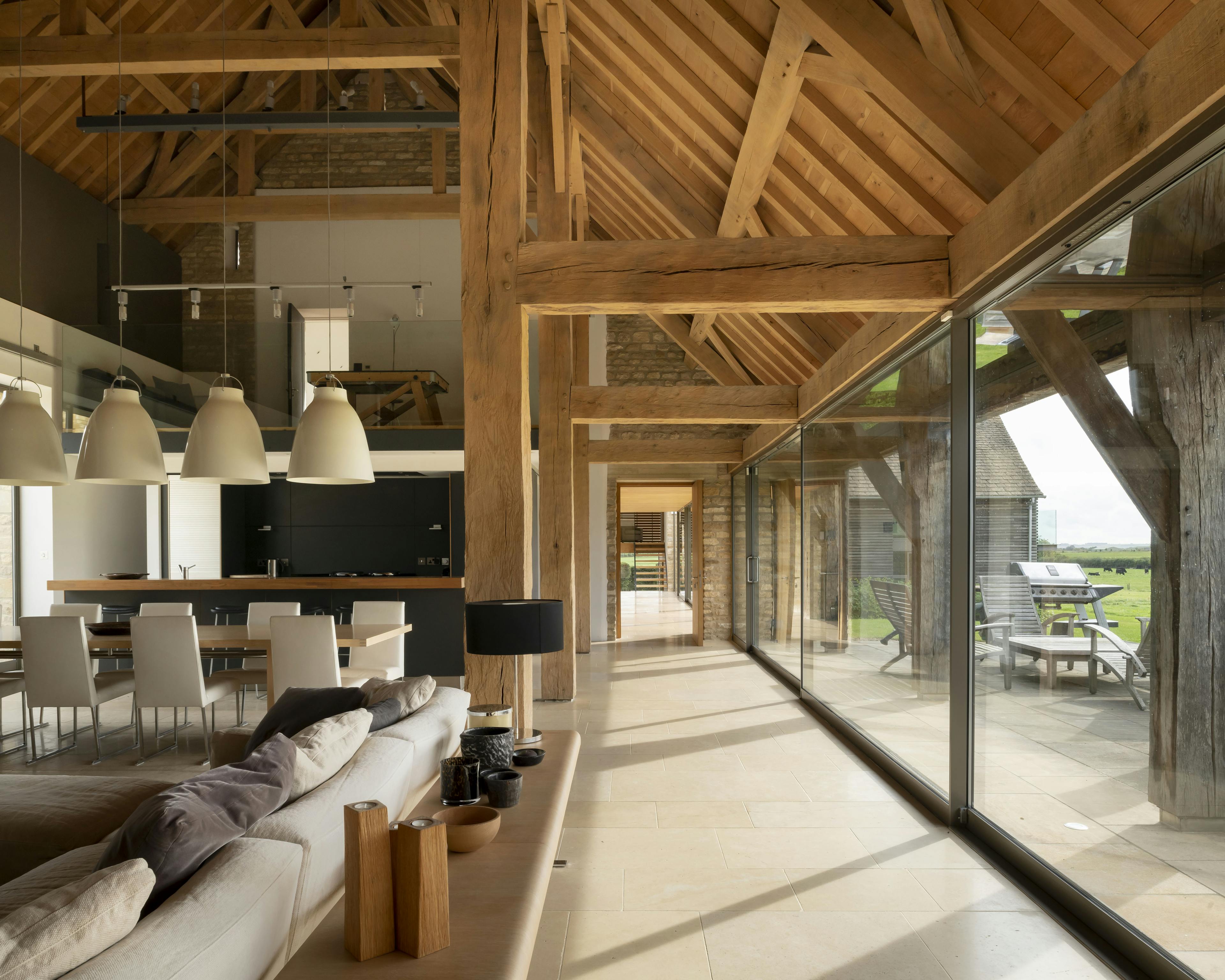 Interior of a renovated oak-framed barn with vaulted ceilings, exposed beams, and a contemporary open-plan living area featuring floor-to-ceiling windows that span the length of the barn and offer sweeping countryside views