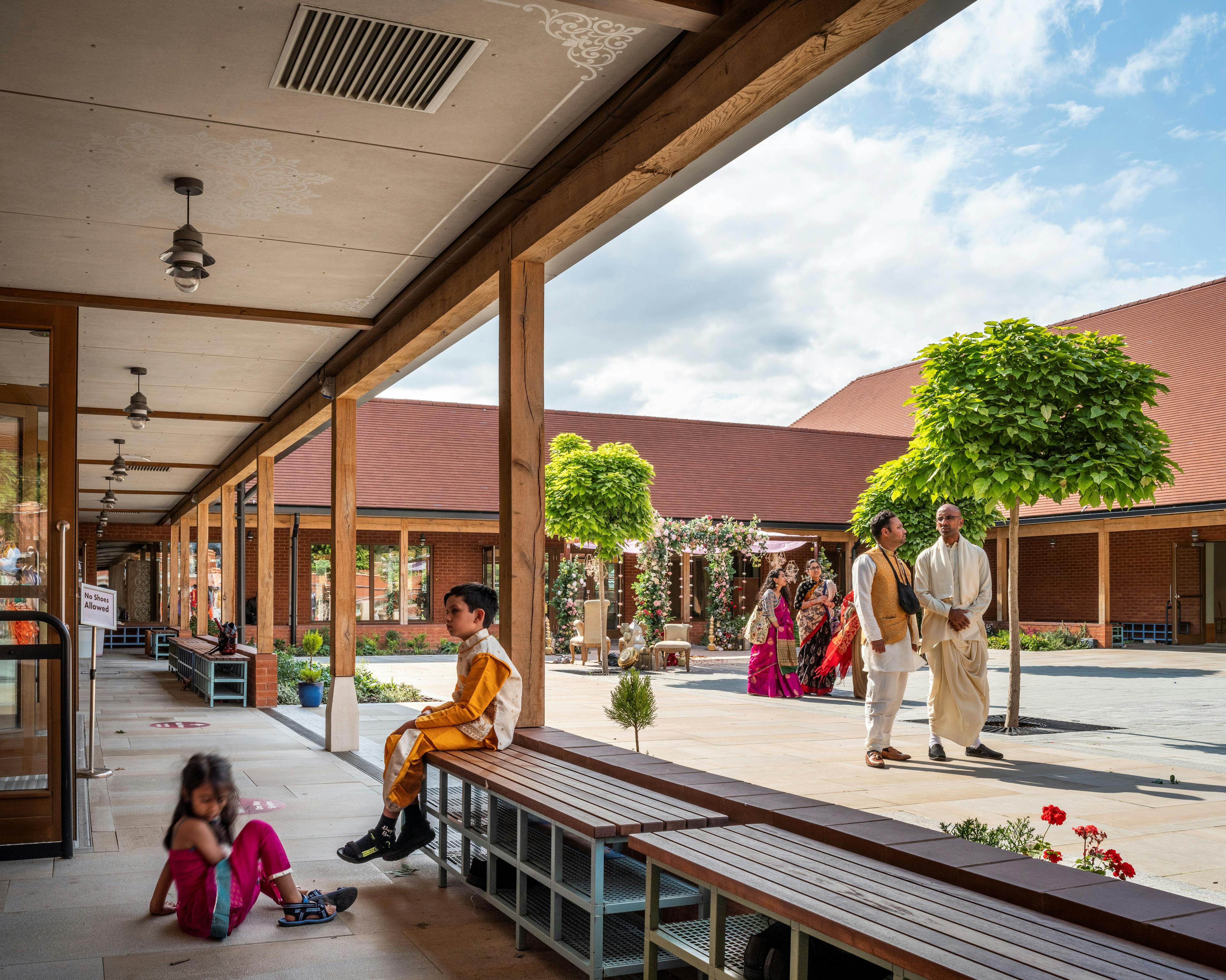 A courtyard with children and adults underneath a green oak veranda of a Hindu Temple
