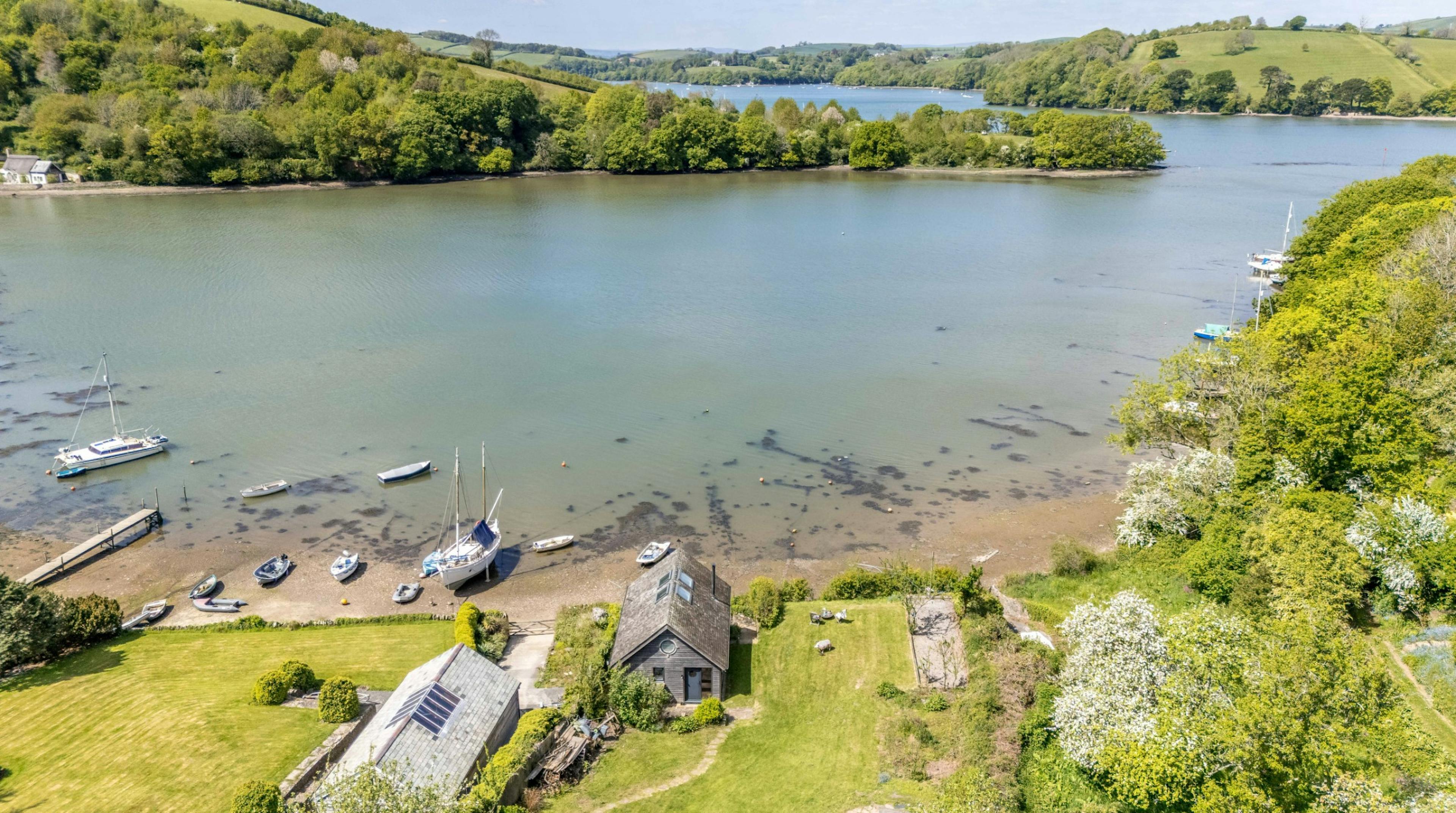 An aerial view of a timber clad boathouse on the edge of a river with view of green hills on the other side of the river