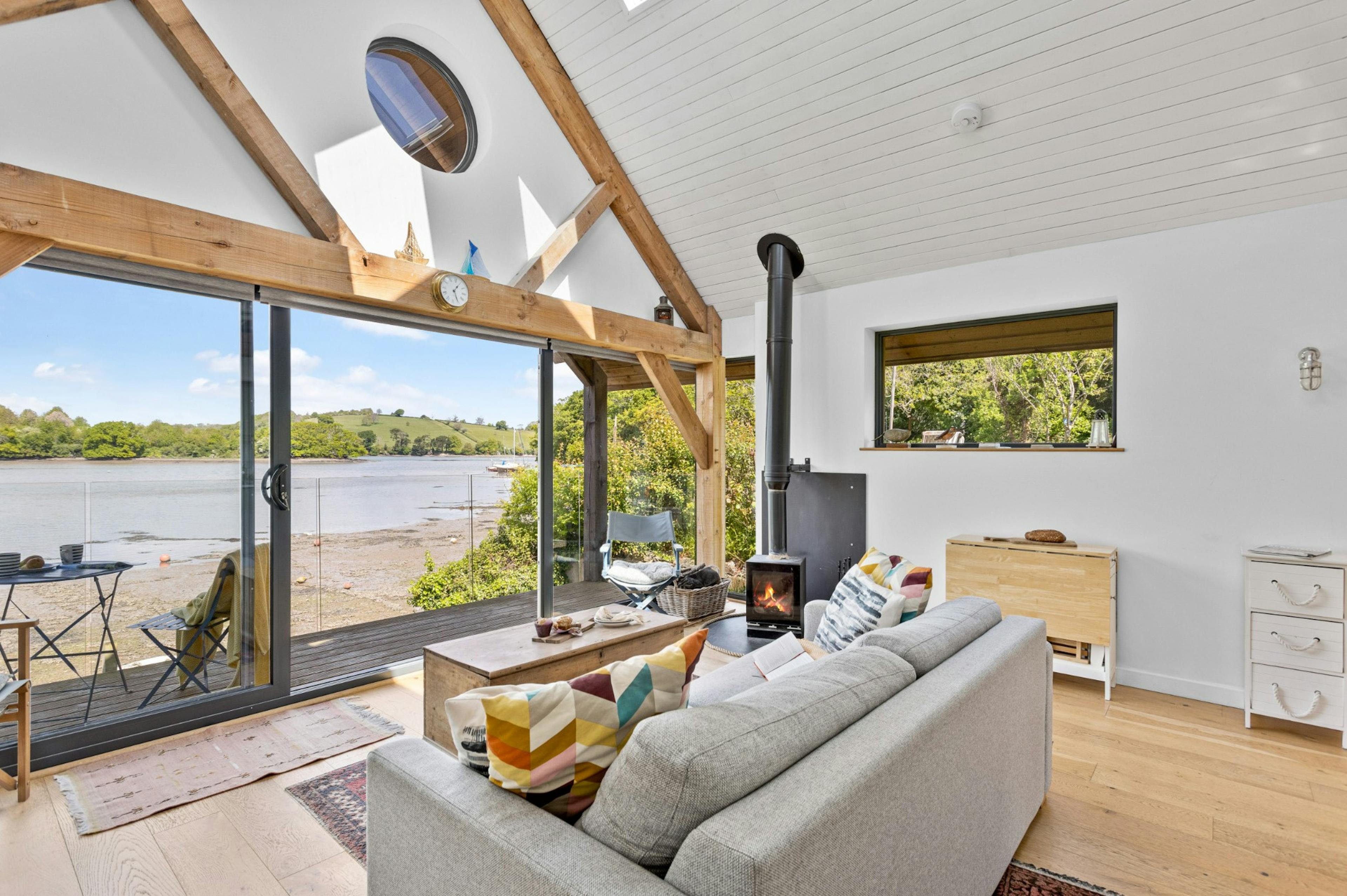 The living room and log burner of an oak framed boathouse on the edge of a river with view of green hills on the other side of the river