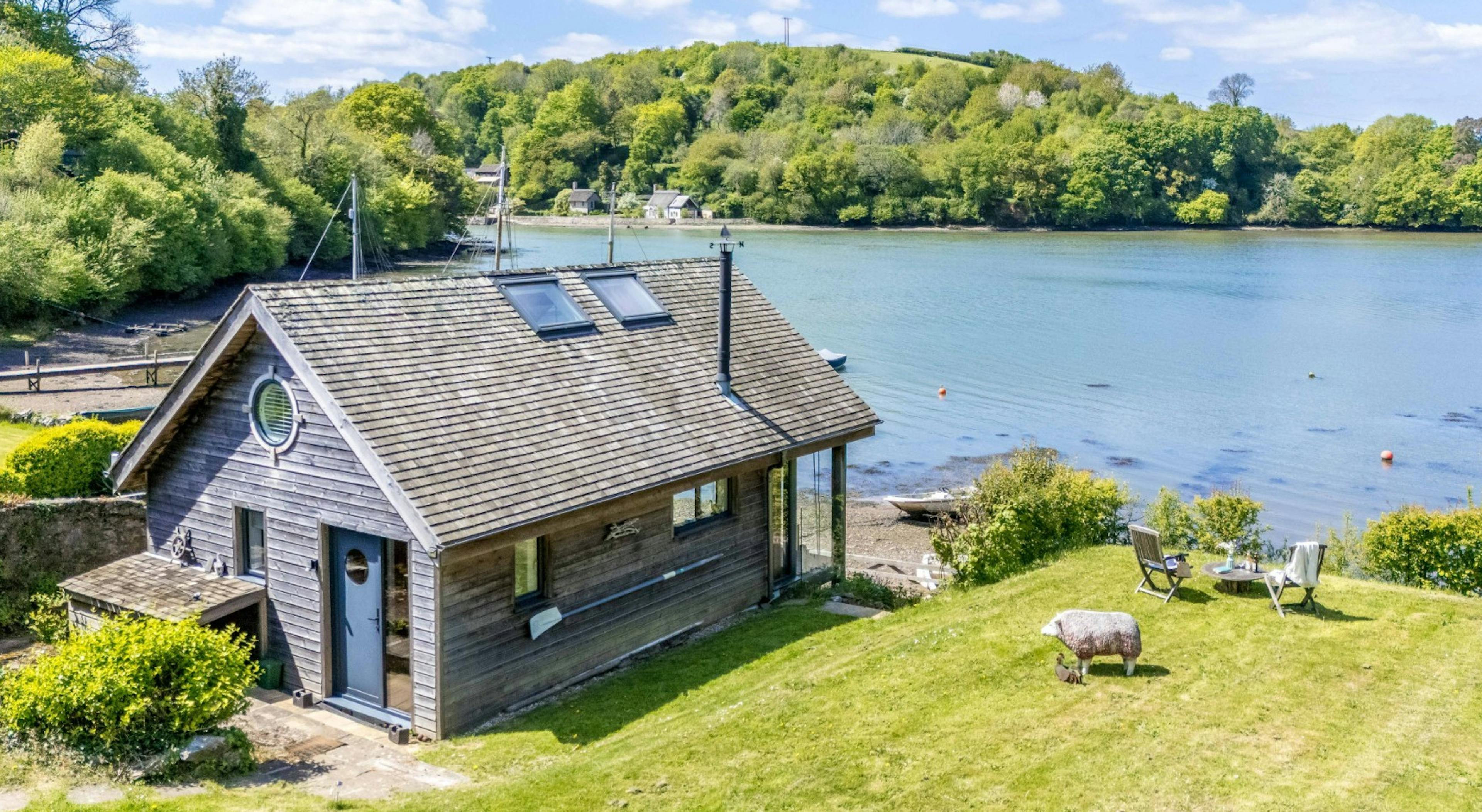 An aerial view of a timber clad boathouse on the edge of a river with view of green hills on the other side of the river