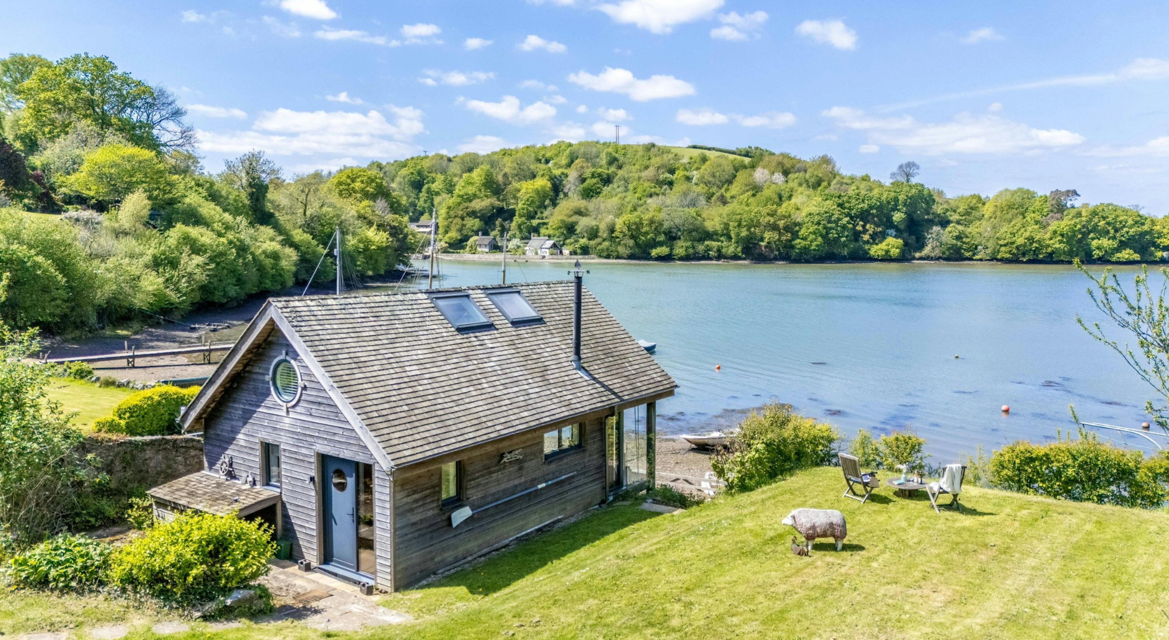 An aerial view of a timber clad boathouse on the edge of a river with view of green hills on the other side of the river