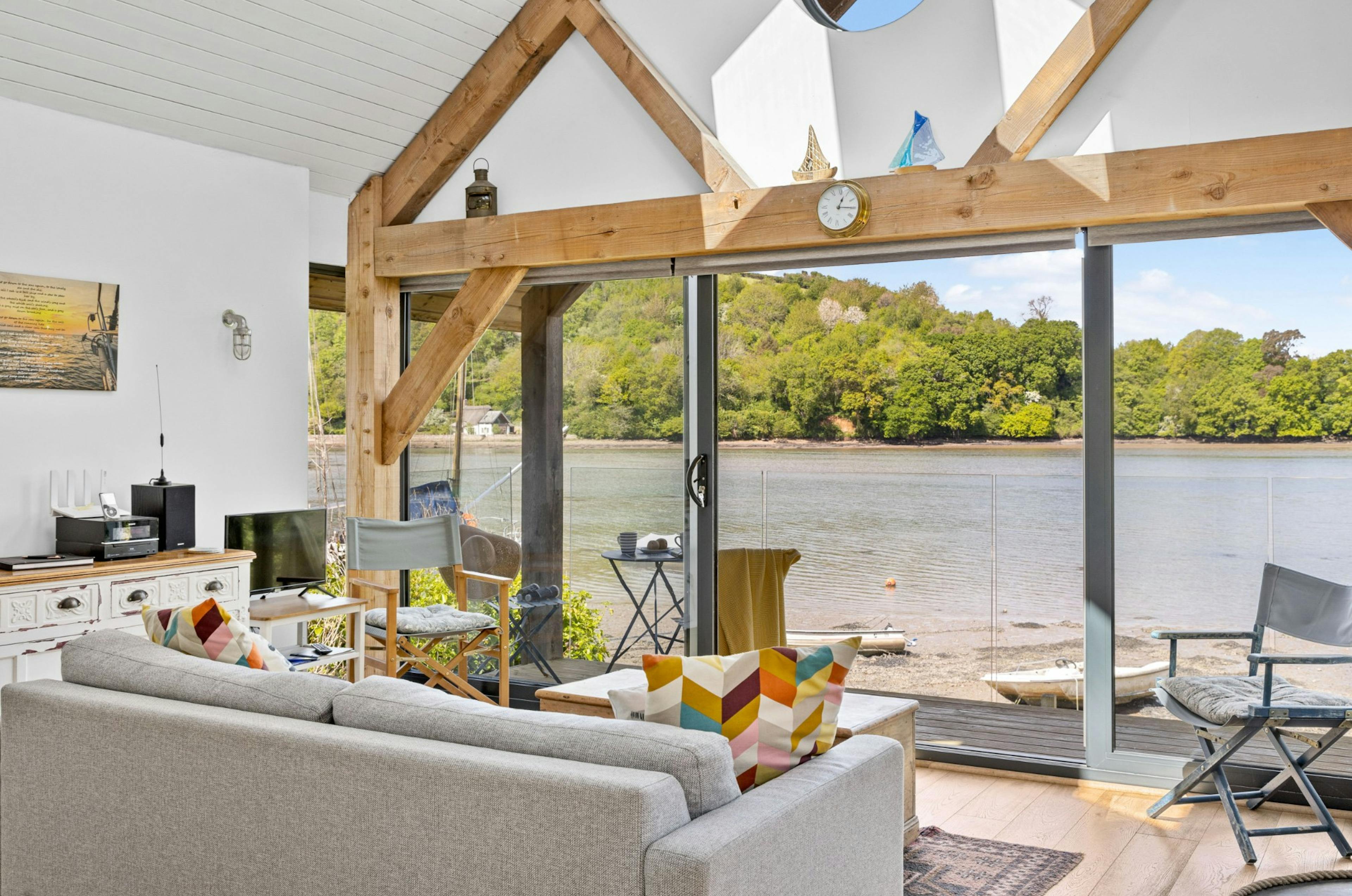 The living room of an oak framed boathouse on the edge of a river with view of green hills on the other side of the river