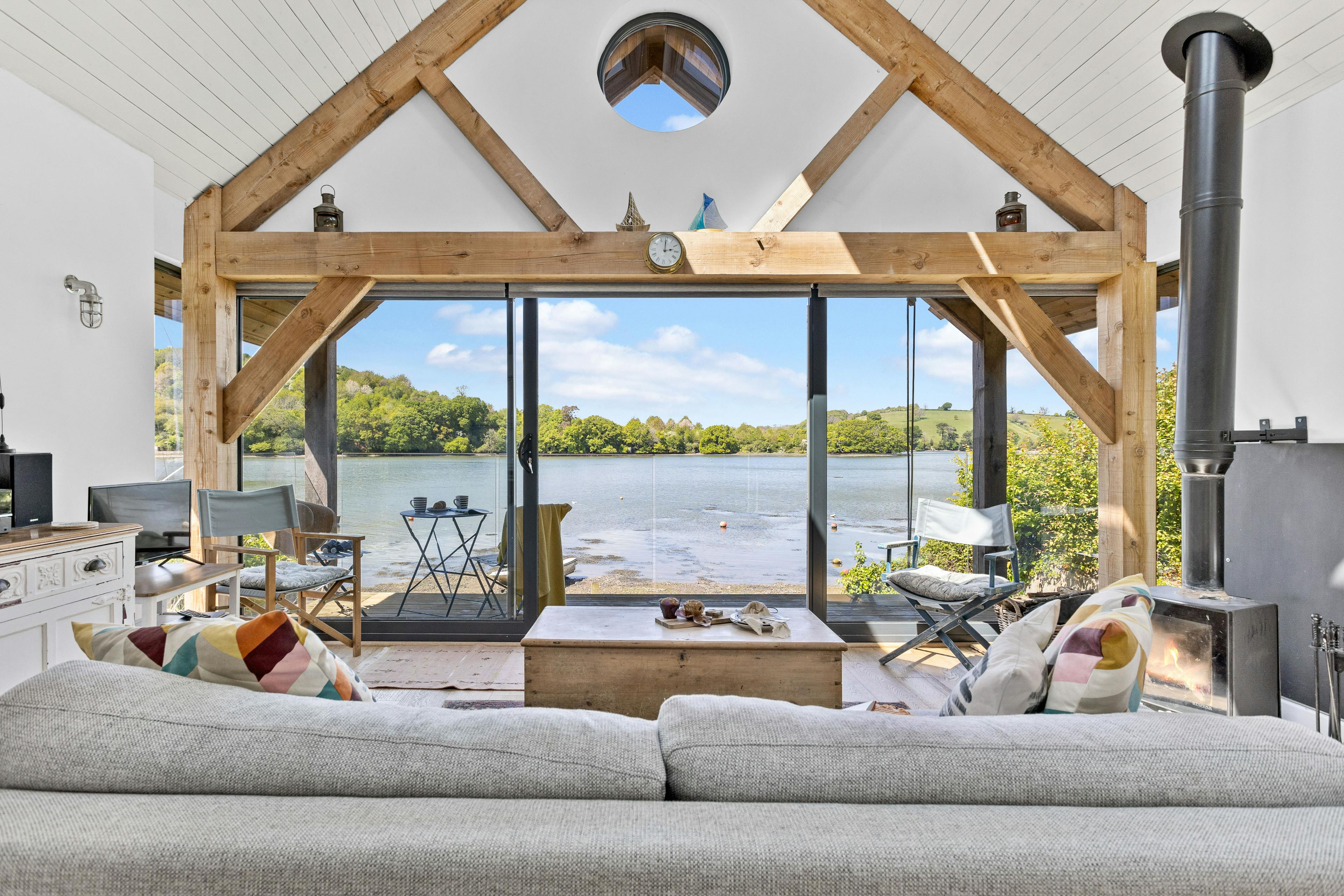 The living room and log burner of an oak framed boathouse on the edge of a river with view of green hills on the other side of the river