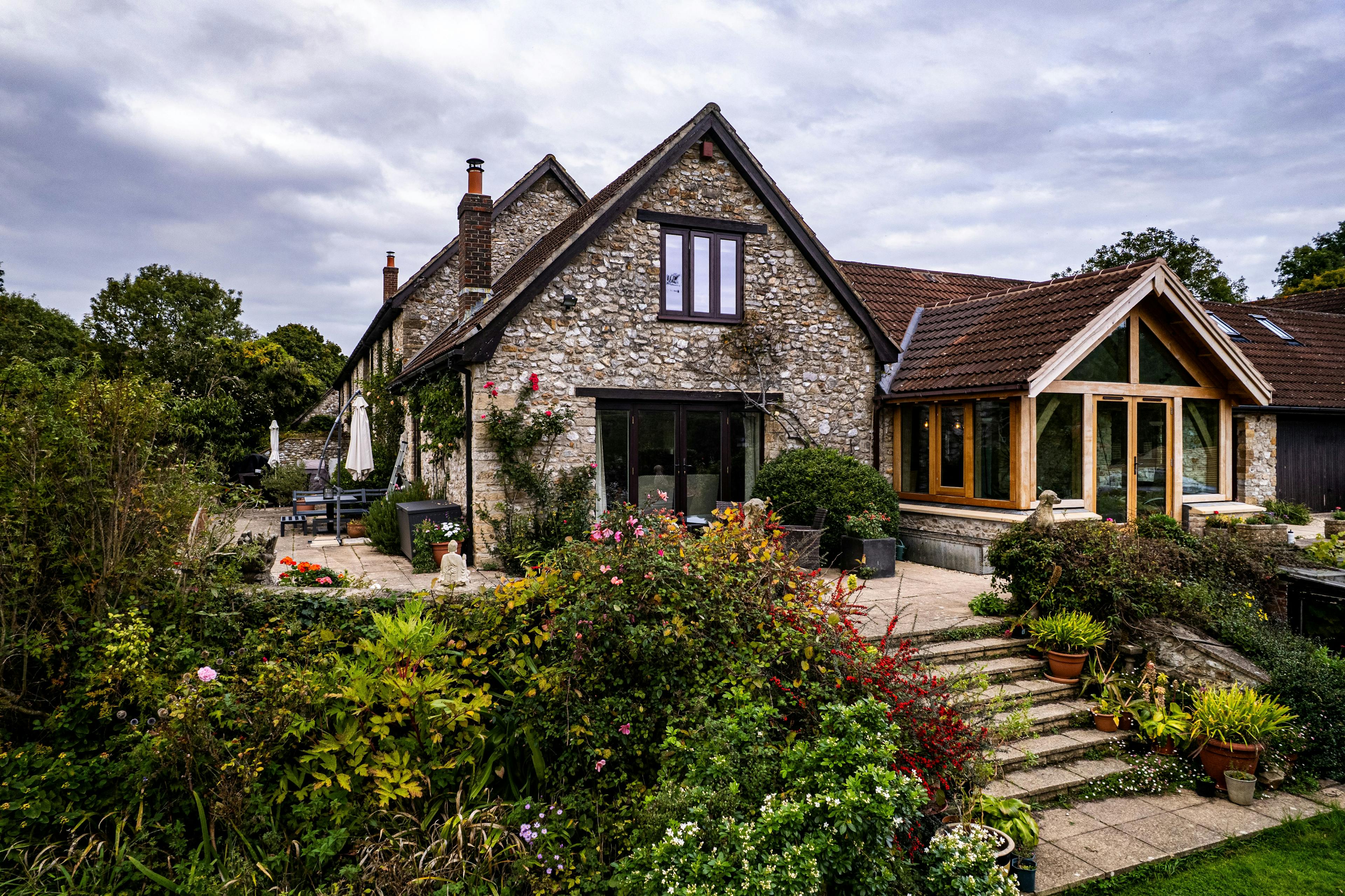 Exterior image of an oak frame garden room extension to a home with natural stone cladding, with direct glazing and situated in a mature garden