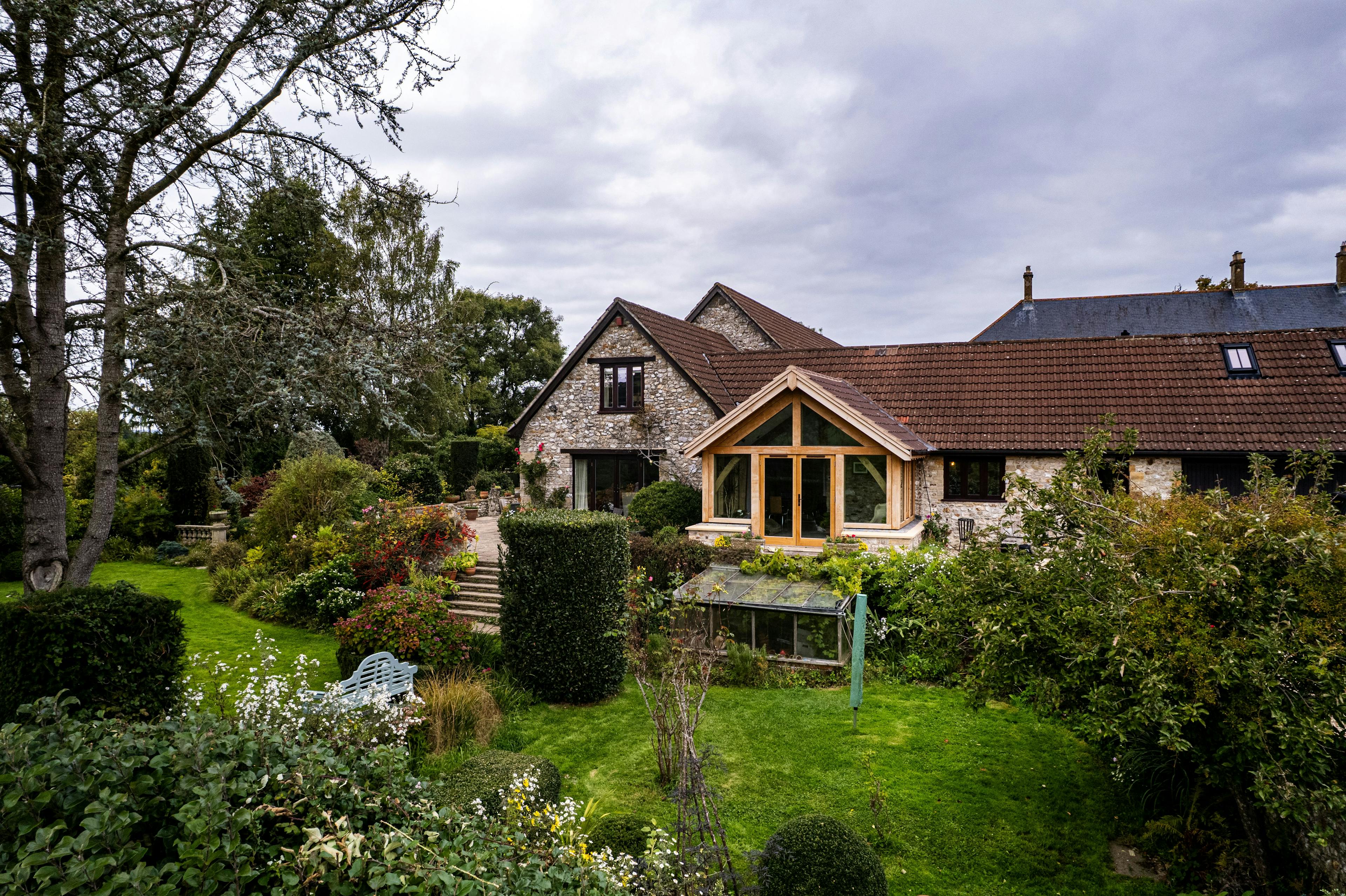 Exterior image of an oak frame garden room extension to a home with natural stone cladding, with direct glazing and situated in a mature garden