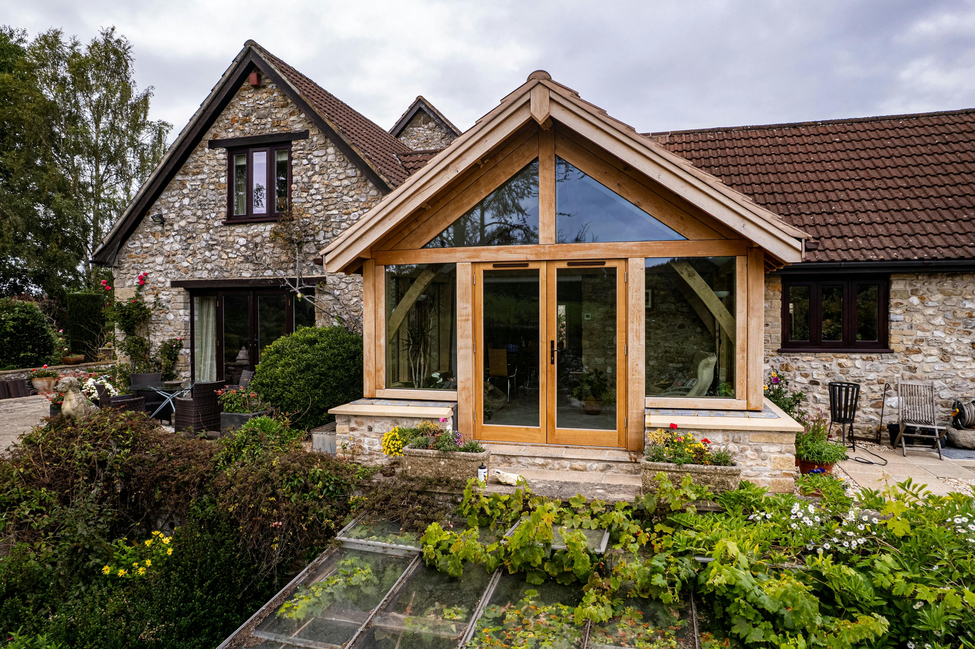 Exterior image of an oak frame garden room extension to a home with natural stone cladding, with direct glazing and situated in a mature garden