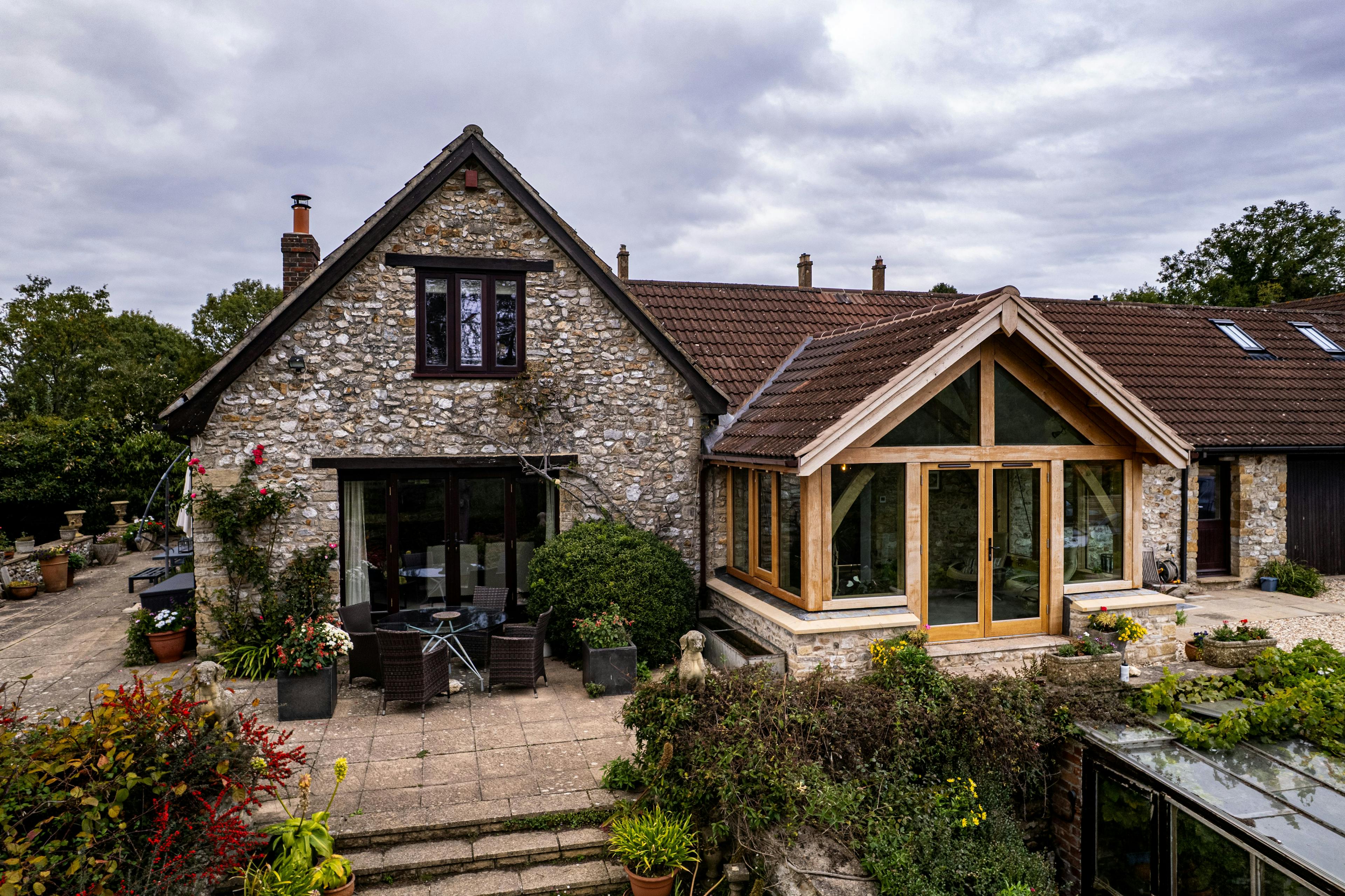 Exterior image of an oak frame garden room extension to a home with natural stone cladding, with direct glazing and situated in a mature garden