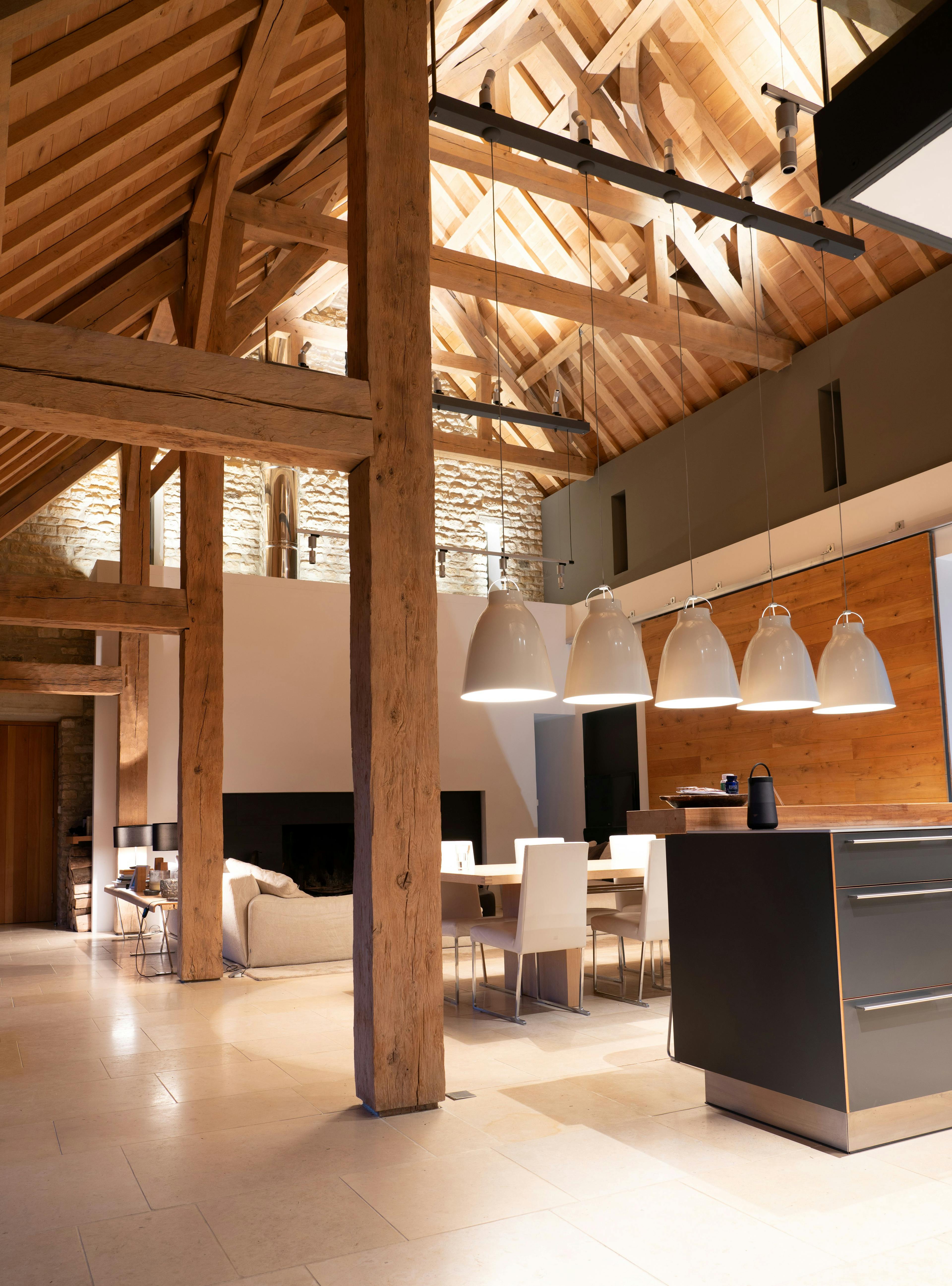 A night time image of an interior of a spacious renovated oak-framed barn featuring a vaulted ceiling, exposed rustic beams, and a contemporary open-plan living, kitchen and dining area