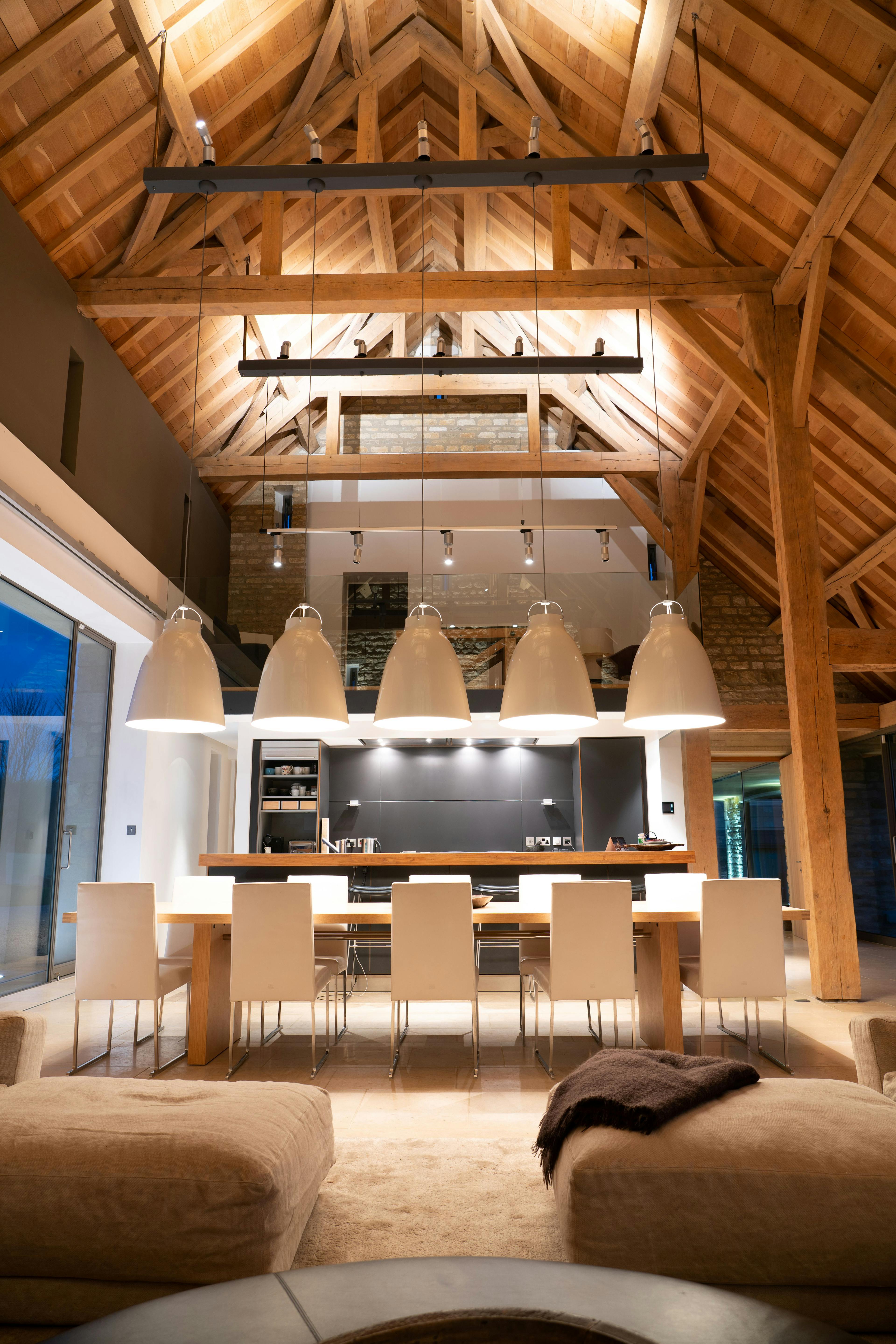A night time image of an interior of a spacious renovated oak-framed barn featuring a vaulted ceiling, exposed rustic beams, and a contemporary open-plan living, kitchen and dining area