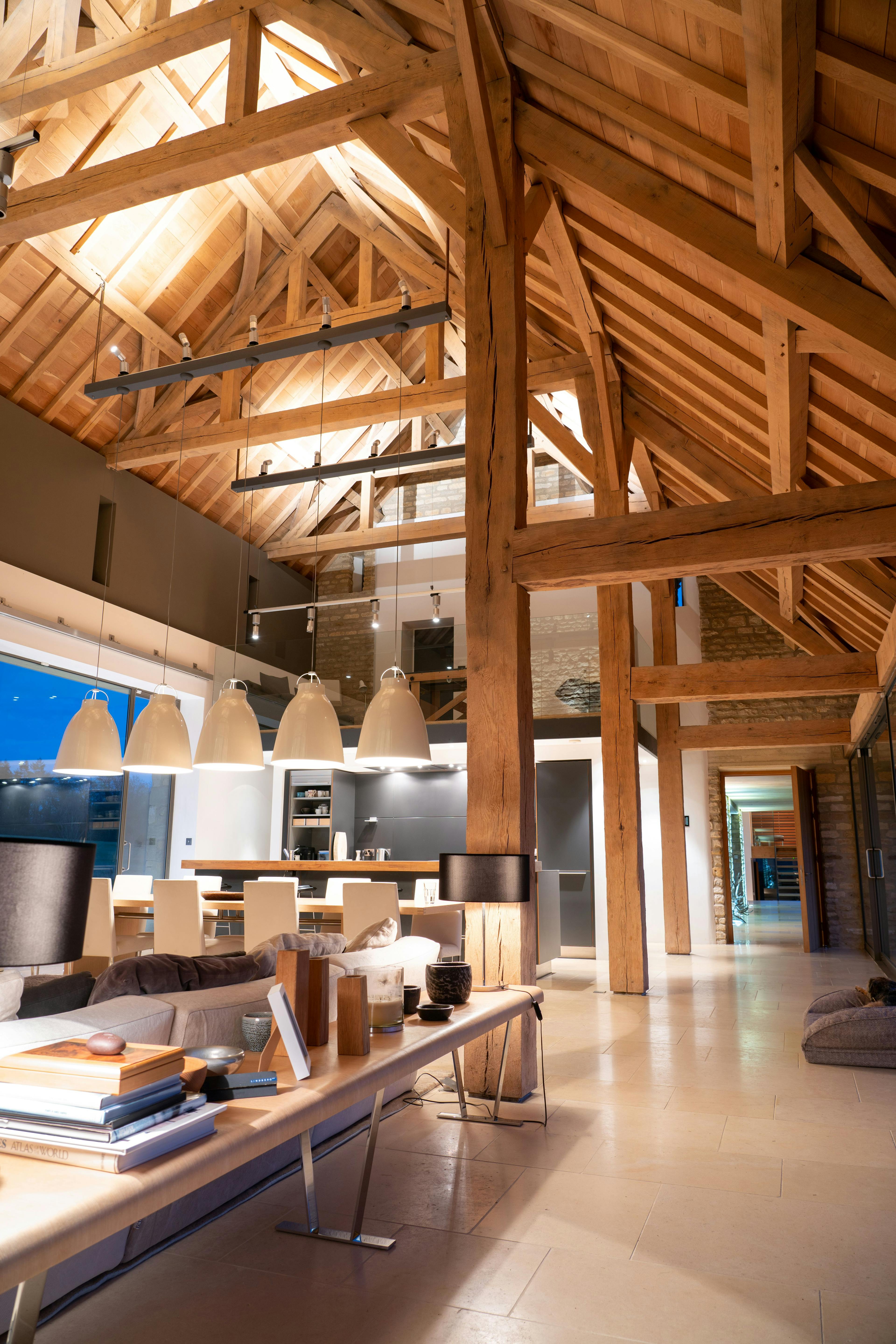 A night time image of an interior of a spacious renovated oak-framed barn featuring a vaulted ceiling, exposed rustic beams, and a contemporary open-plan living, kitchen and dining area