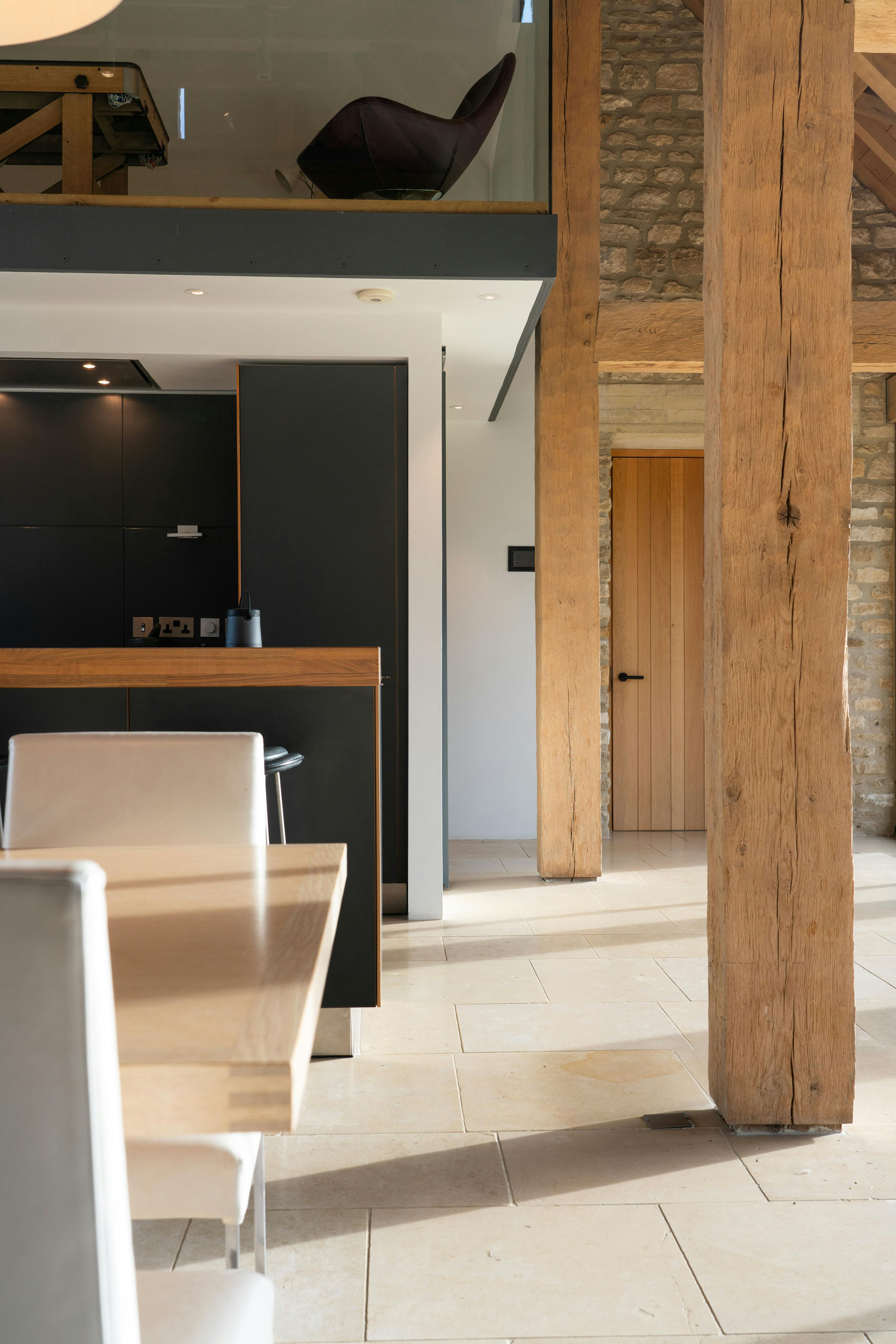 Sunlight beams onto an interior of a spacious renovated oak-framed barn featuring exposed rustic beams and a contemporary open-plan dining area and kitchen