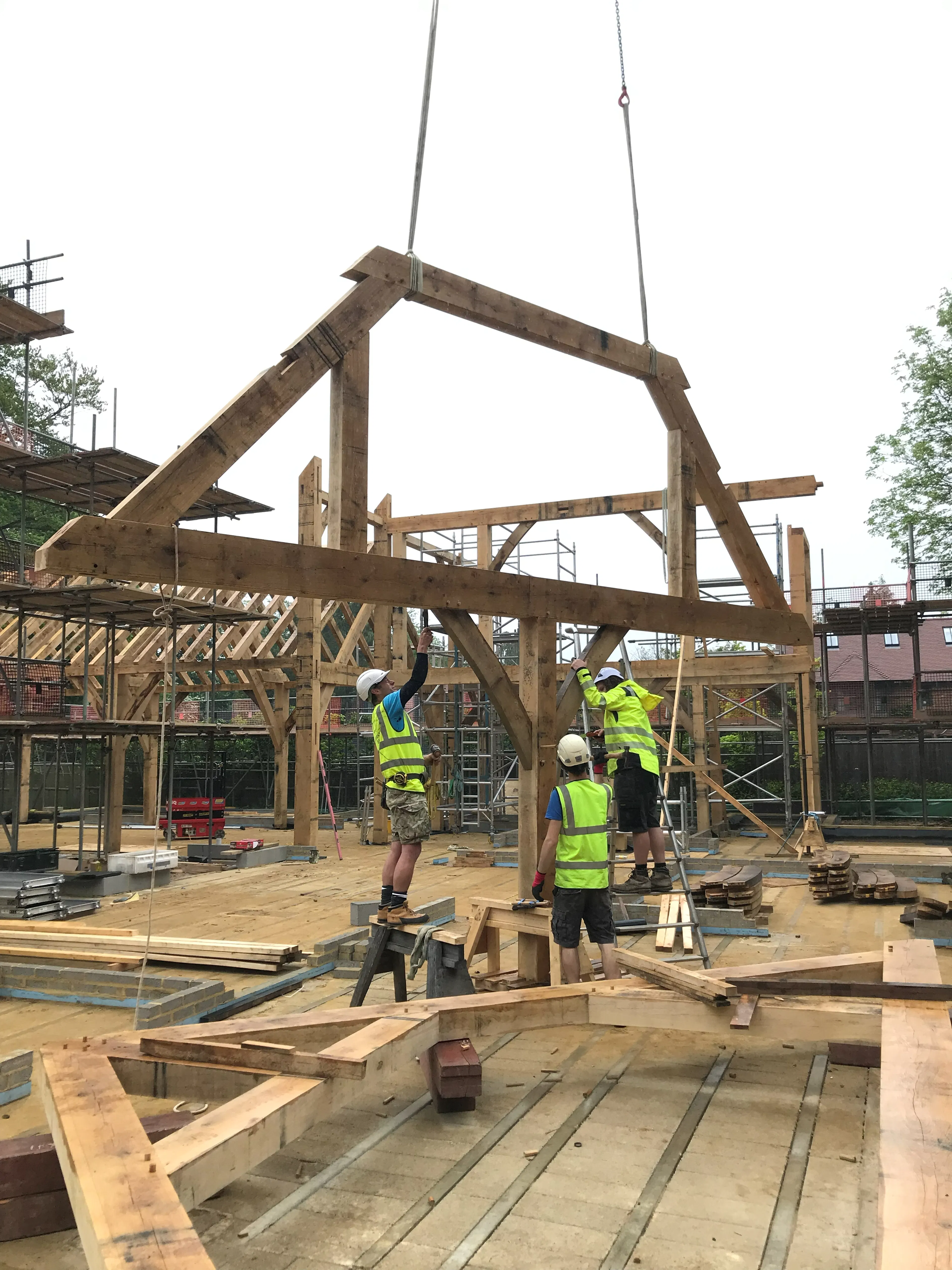 Carpenters monitor a crane as it lowers a truss into place during installation of a large 6 bedroom oak framed family home