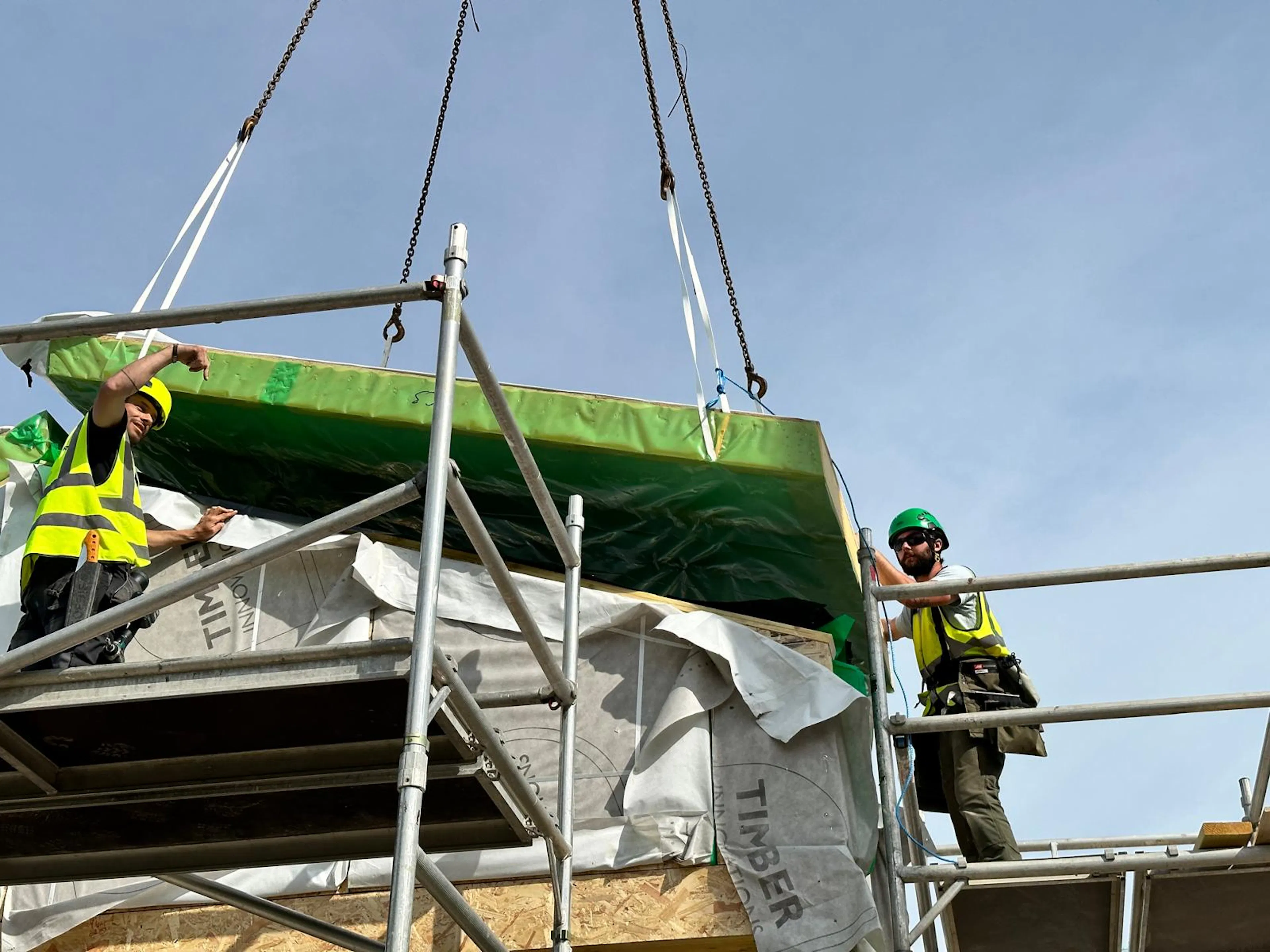 Two carpenters installing prefabricated panels into a structural oak timber frame at a building site, working on interior wall assembly